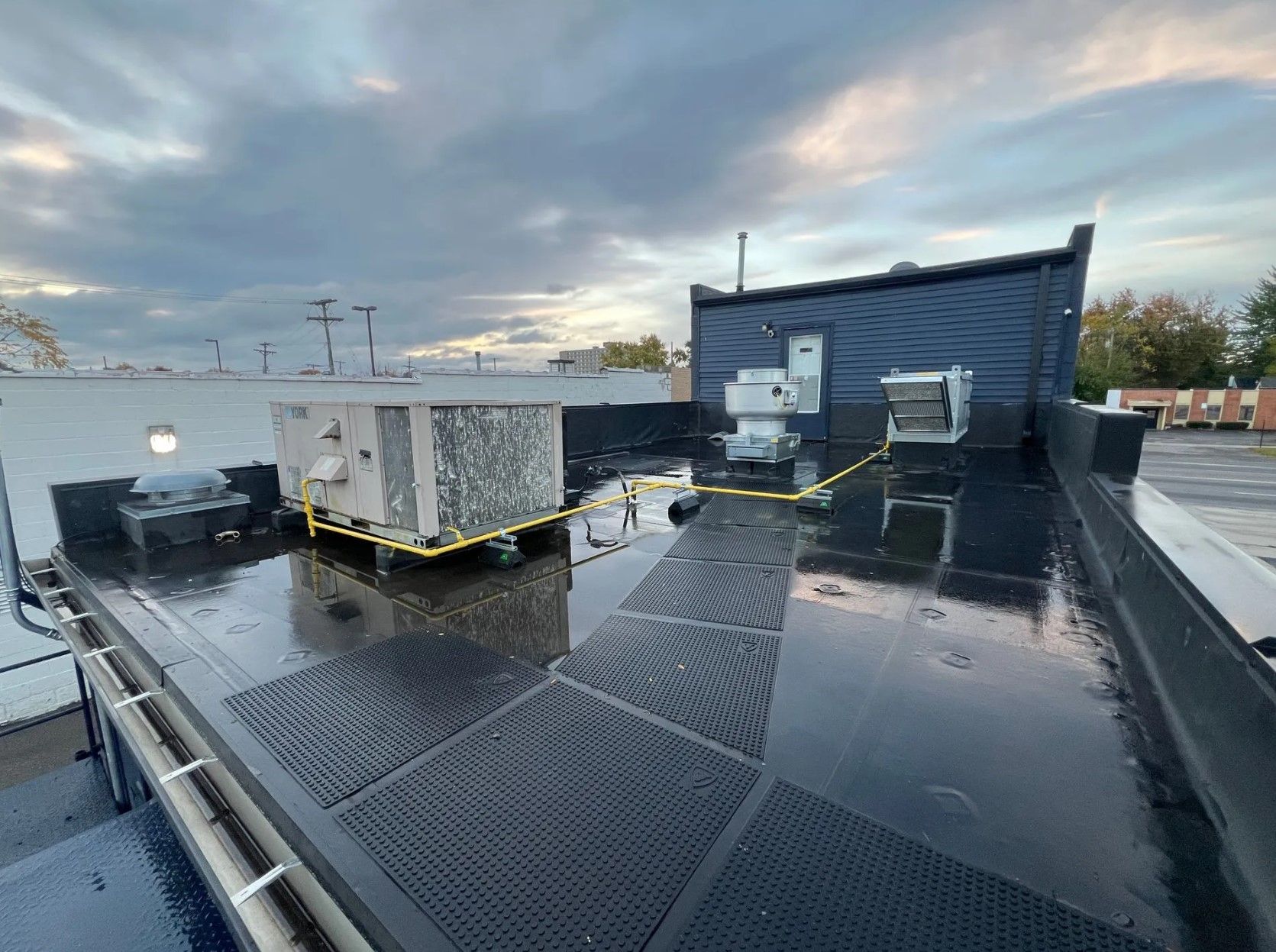 View of a flat, wet commercial rooftop with HVAC units. Dark roofing with safety lines and mats. Overcast sky.