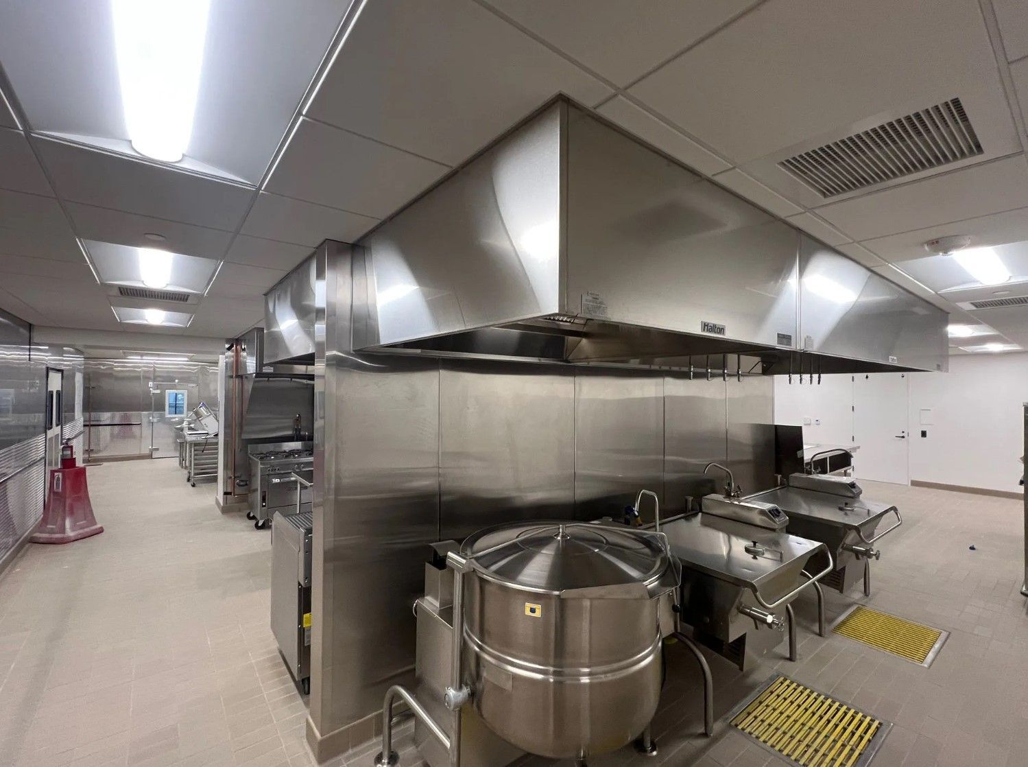 Commercial kitchen with stainless steel equipment and exhaust hoods, overhead lighting, beige floor.