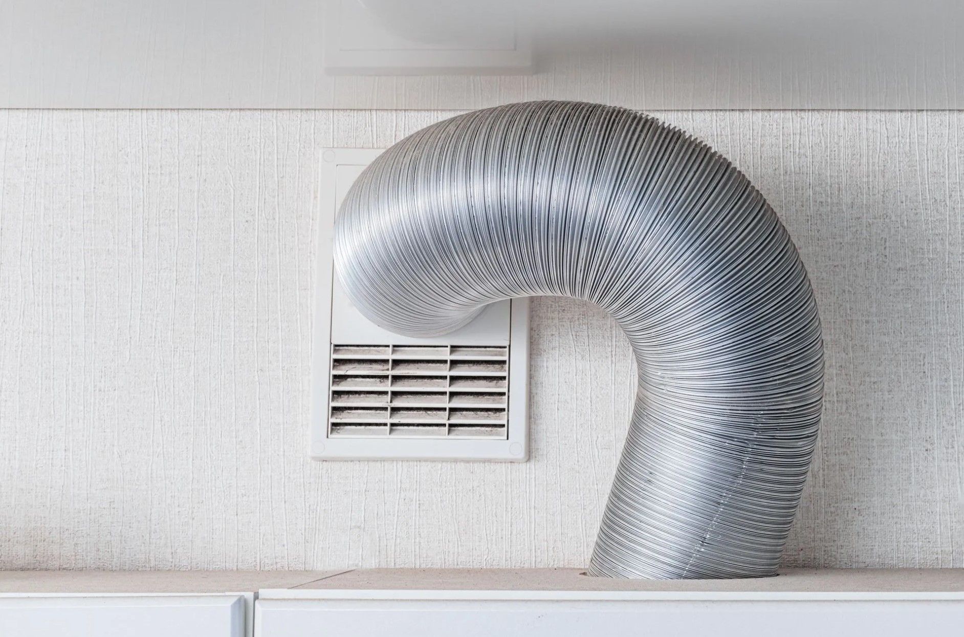 Flexible silver ducting connected to a white rectangular vent on a textured white wall.