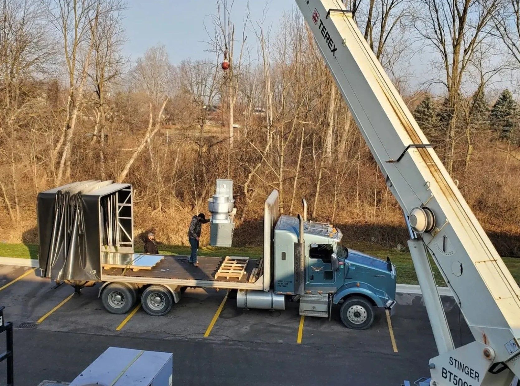 A crane lifting equipment onto a flatbed truck; a worker stands nearby.