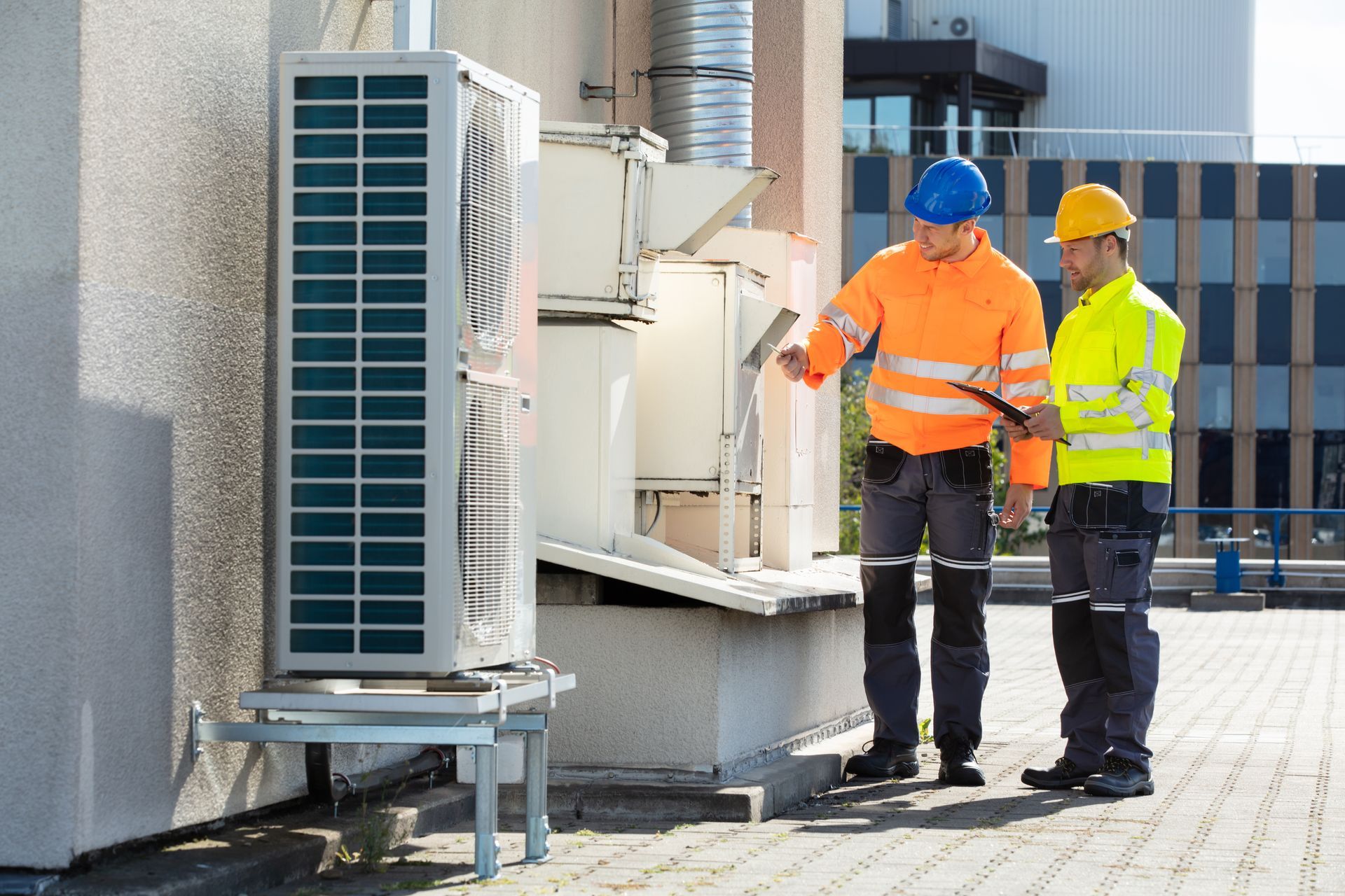Two HVAC technicians examine an outdoor air conditioning unit on a building rooftop.