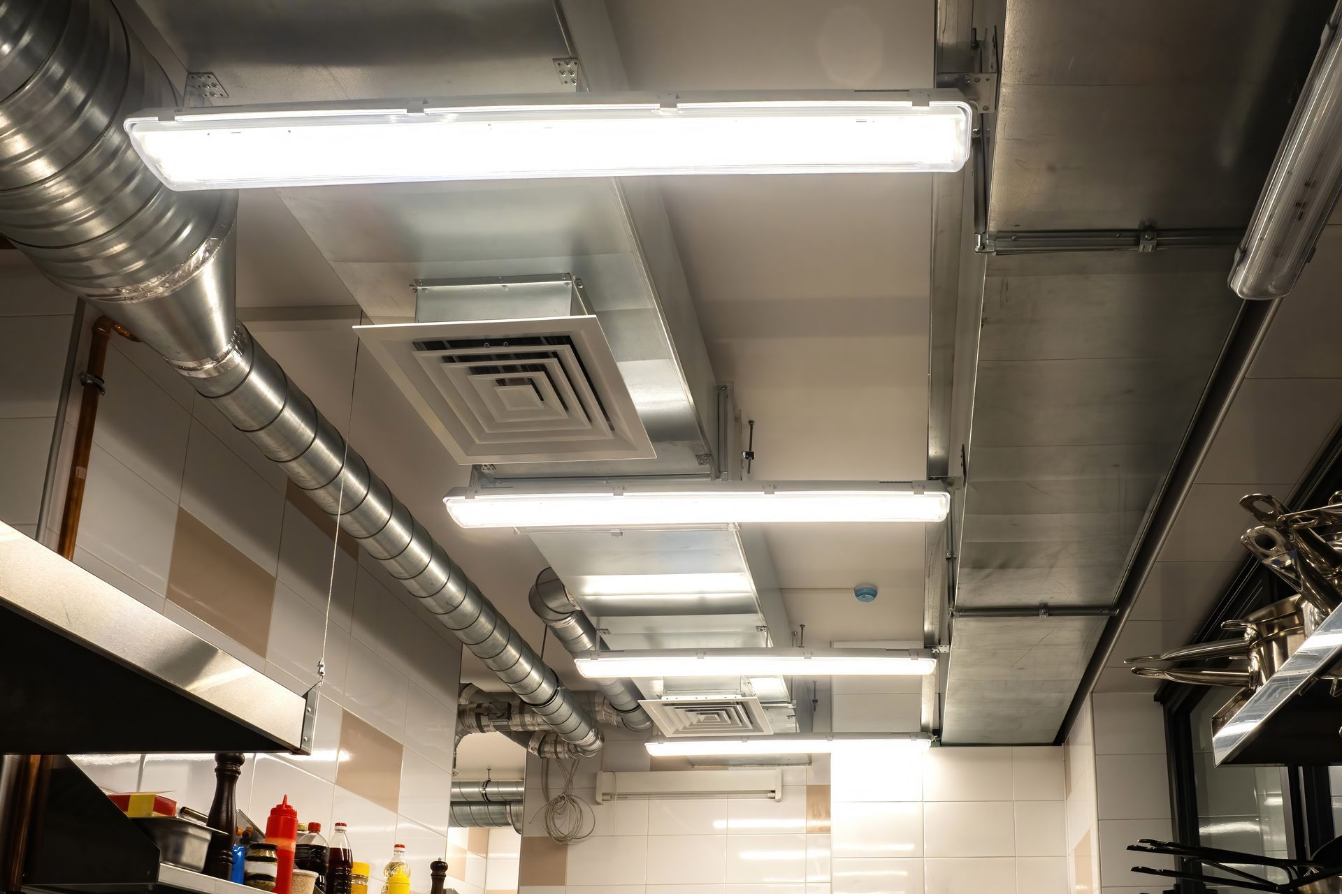 Ceiling with ventilation ducts, fluorescent lights, and a vent in a commercial kitchen.