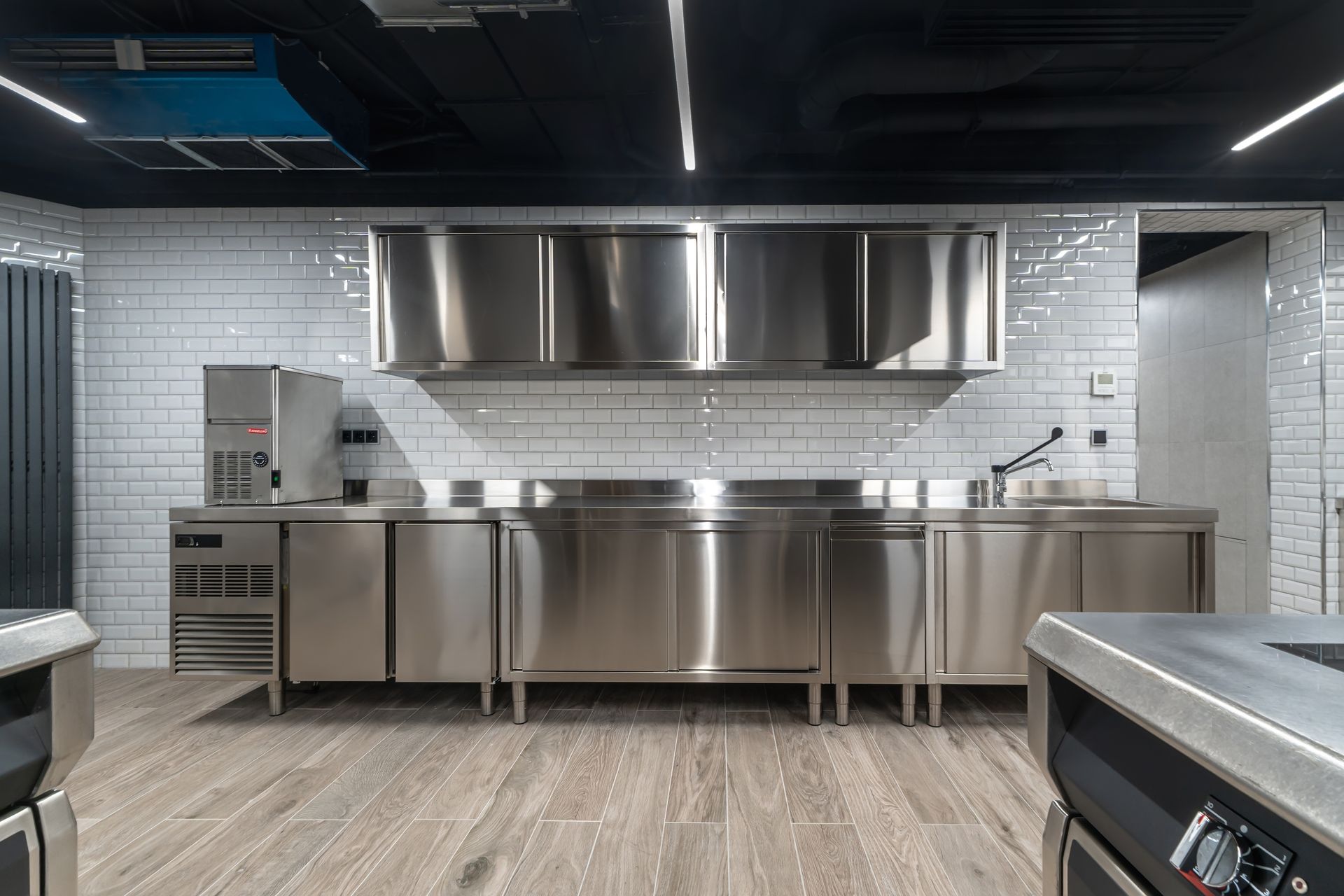 Stainless steel commercial kitchen with vent hood, counters, and appliances. White brick wall, dark ceiling.