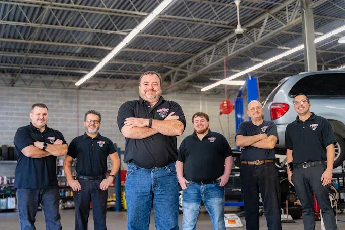 Six men in black shirts in a car repair shop; arms crossed, posing. A car is in the background.