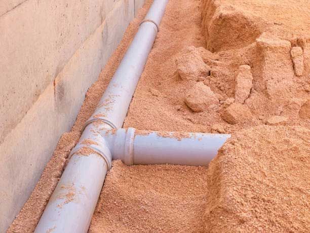 Gray PVC pipes in a trench filled with sawdust, near a concrete wall.