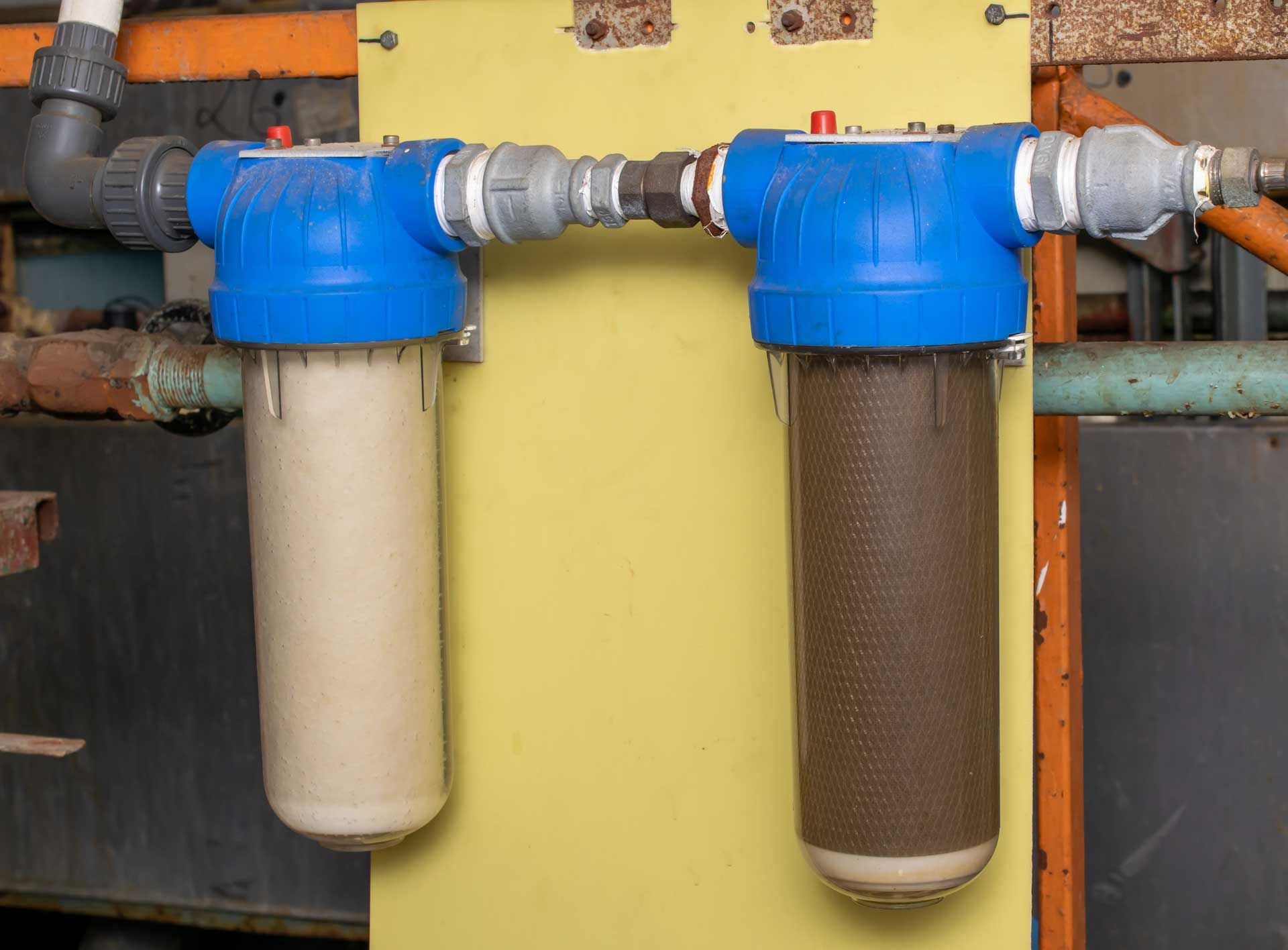 Two water filters; one clean, one brown and dirty, connected by pipes, mounted on a yellow wall.