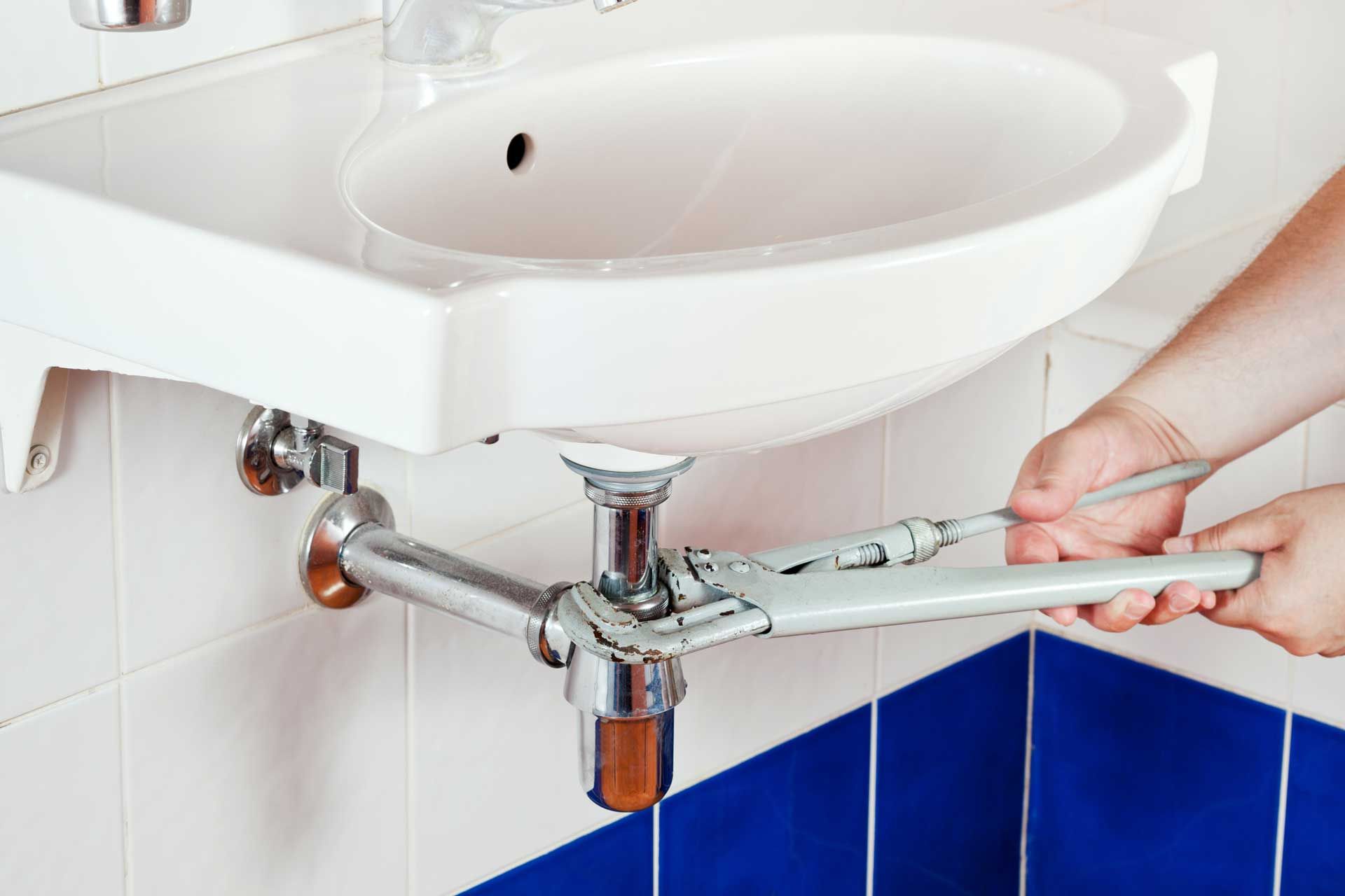 Plumber fixing a sink drain with a wrench in a bathroom with blue tile.