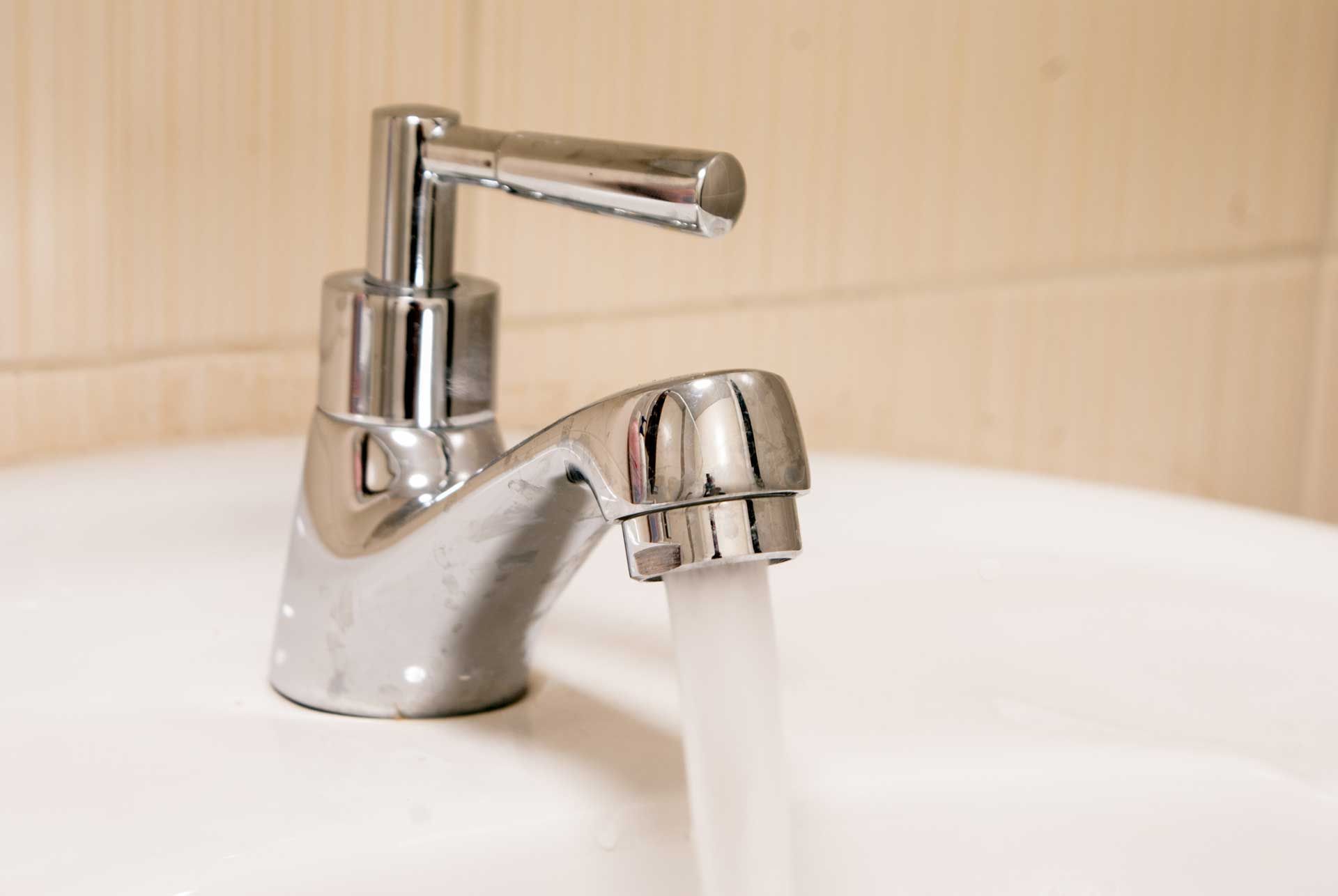 Chrome faucet with running water in a white sink, against a light-colored tile wall.