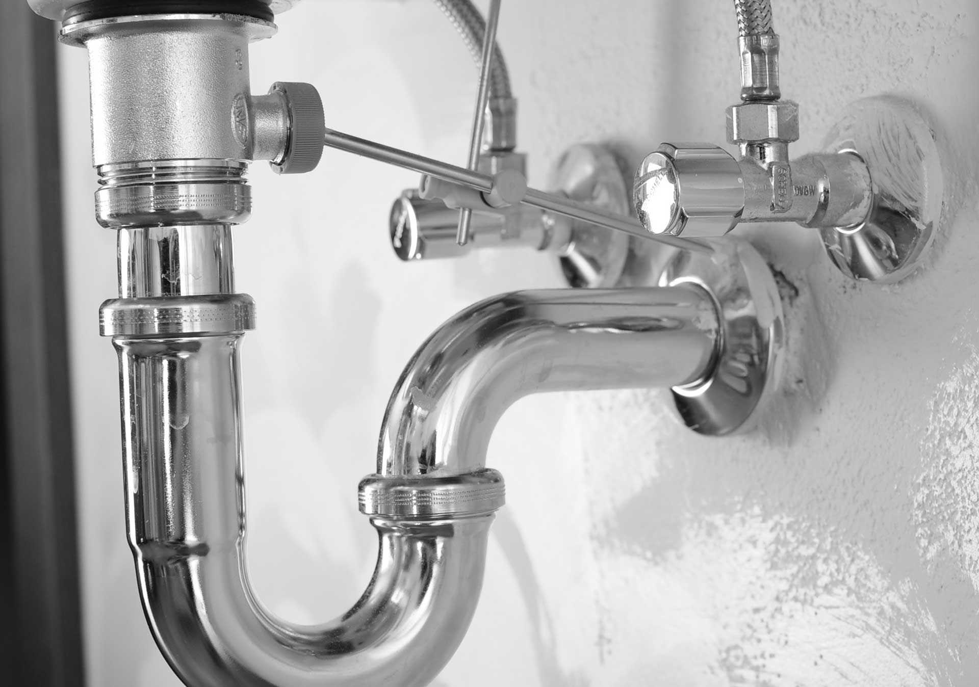 Shiny plumbing under a sink: drain pipe, trap, and water valves mounted on a white wall.