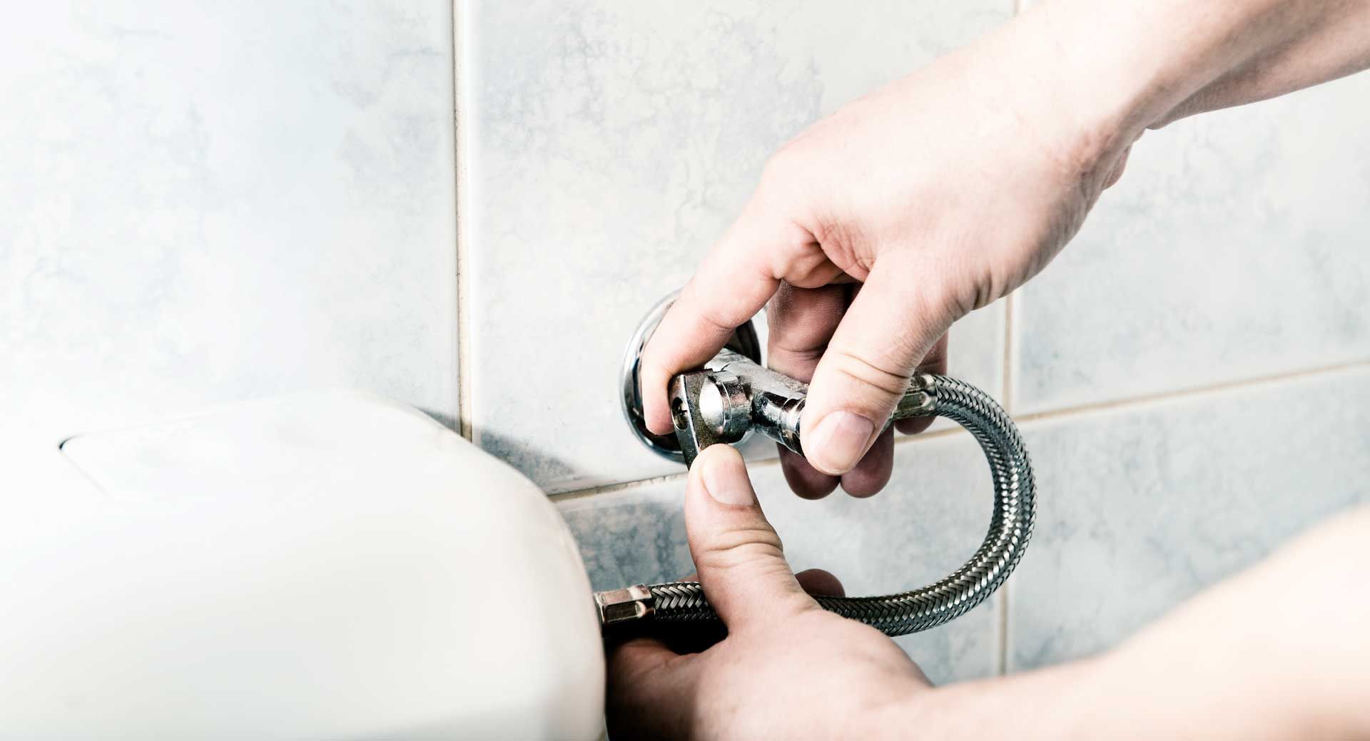 Hands tightening a flexible water supply line to a toilet, near tiled wall.