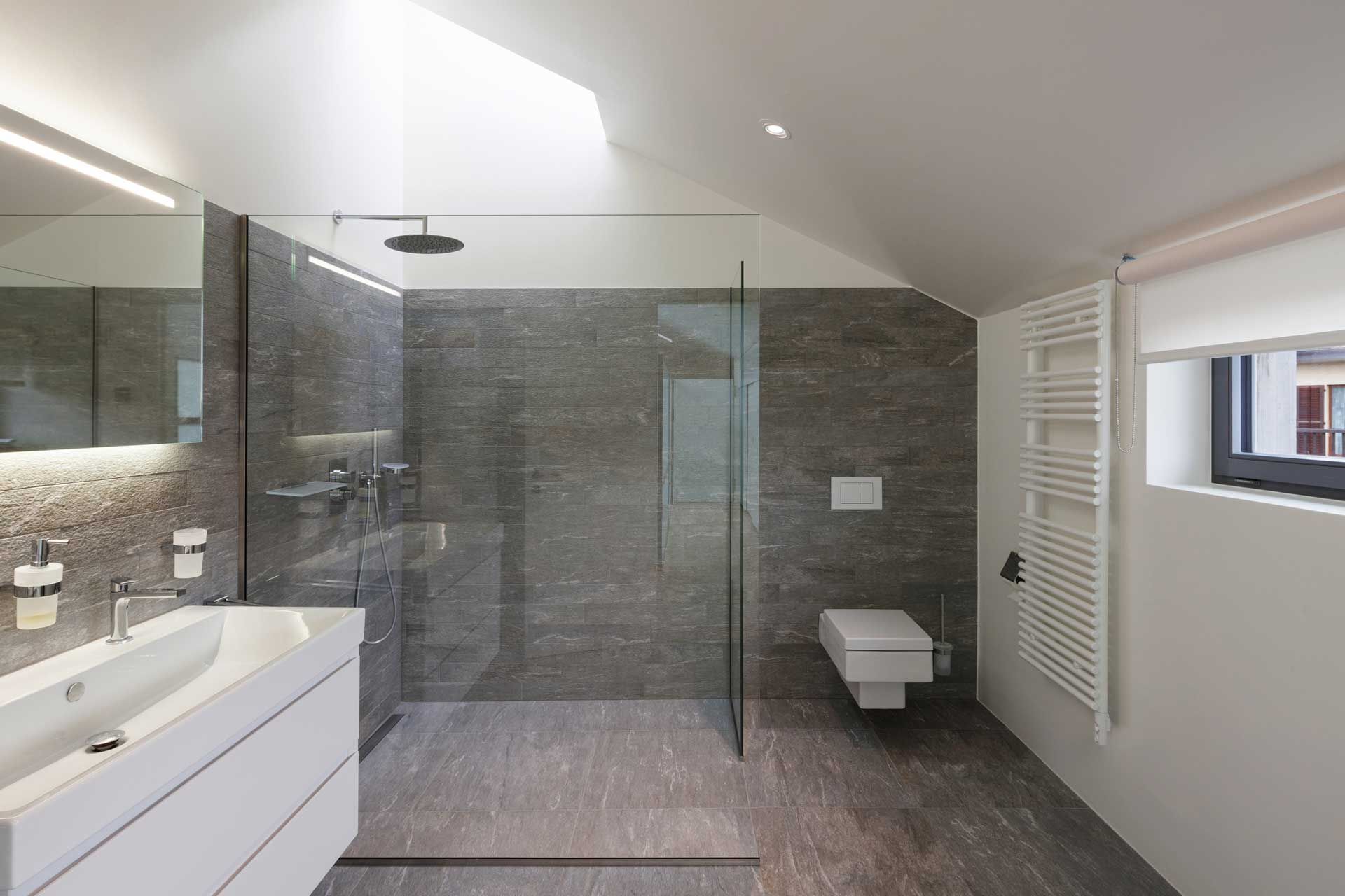 Modern bathroom with gray tiled shower, white sink and toilet, and a window.