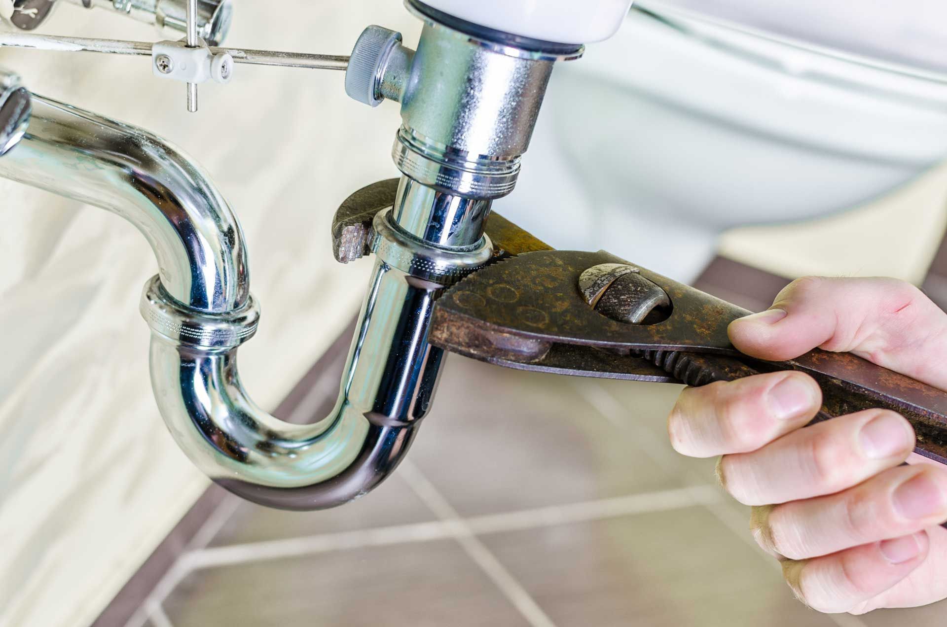 Plumber's hand uses wrench to tighten a chrome pipe under a sink.