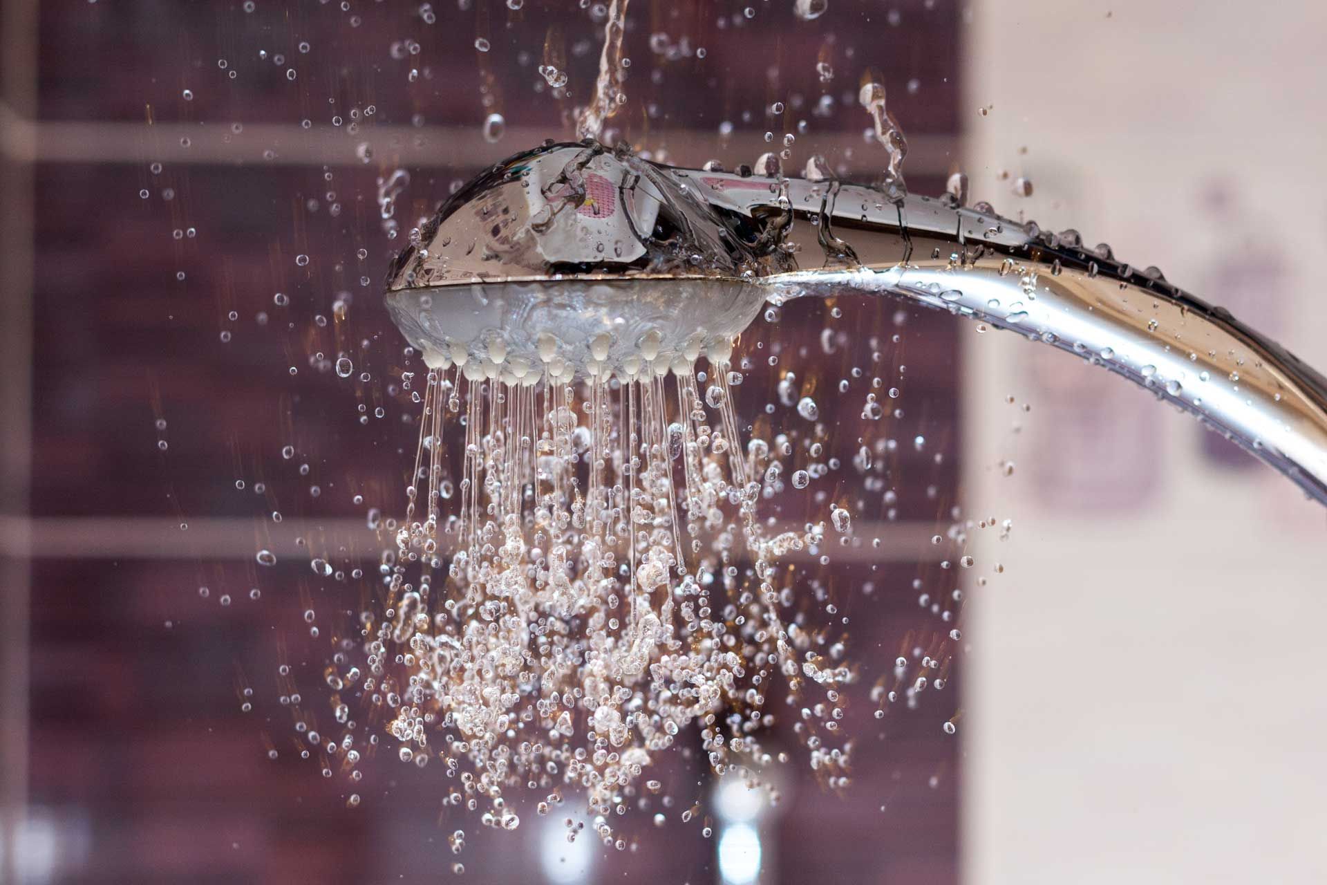 Showerhead with water pouring down, spraying droplets. Chrome fixture against tiled wall.