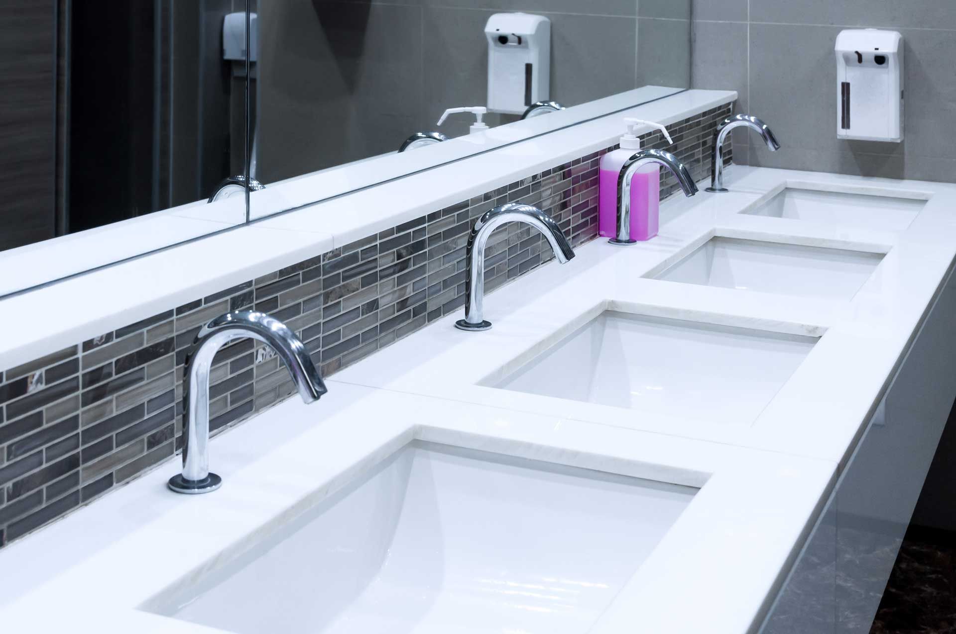 Bathroom with three sinks, chrome faucets, and soap dispensers, with a mirror above.