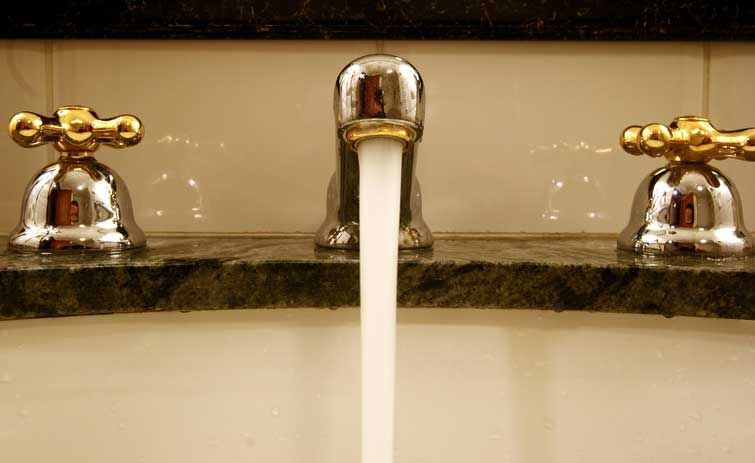 Faucet with running water, flanked by two ornate chrome and gold-colored handles, set on a dark marble counter.