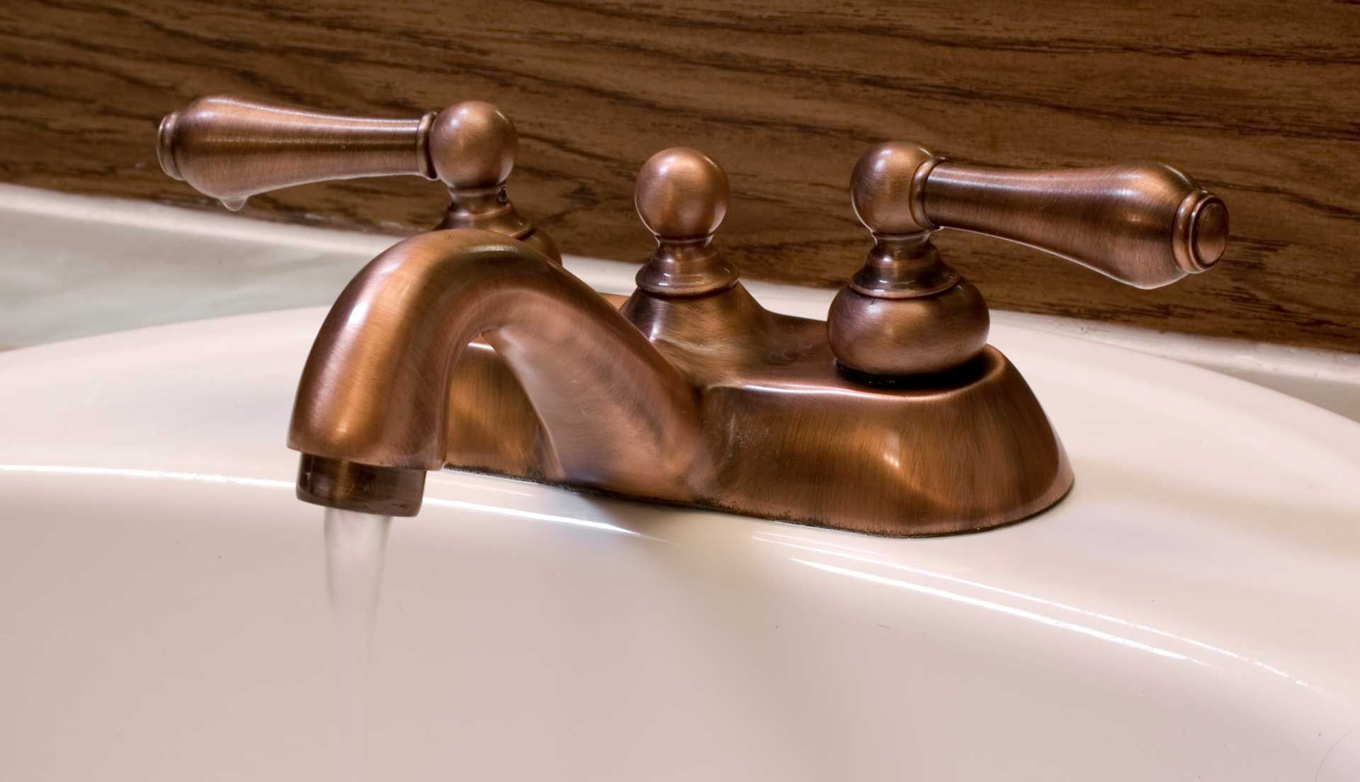 Bronze faucet with running water in a white sink, against a wooden background.