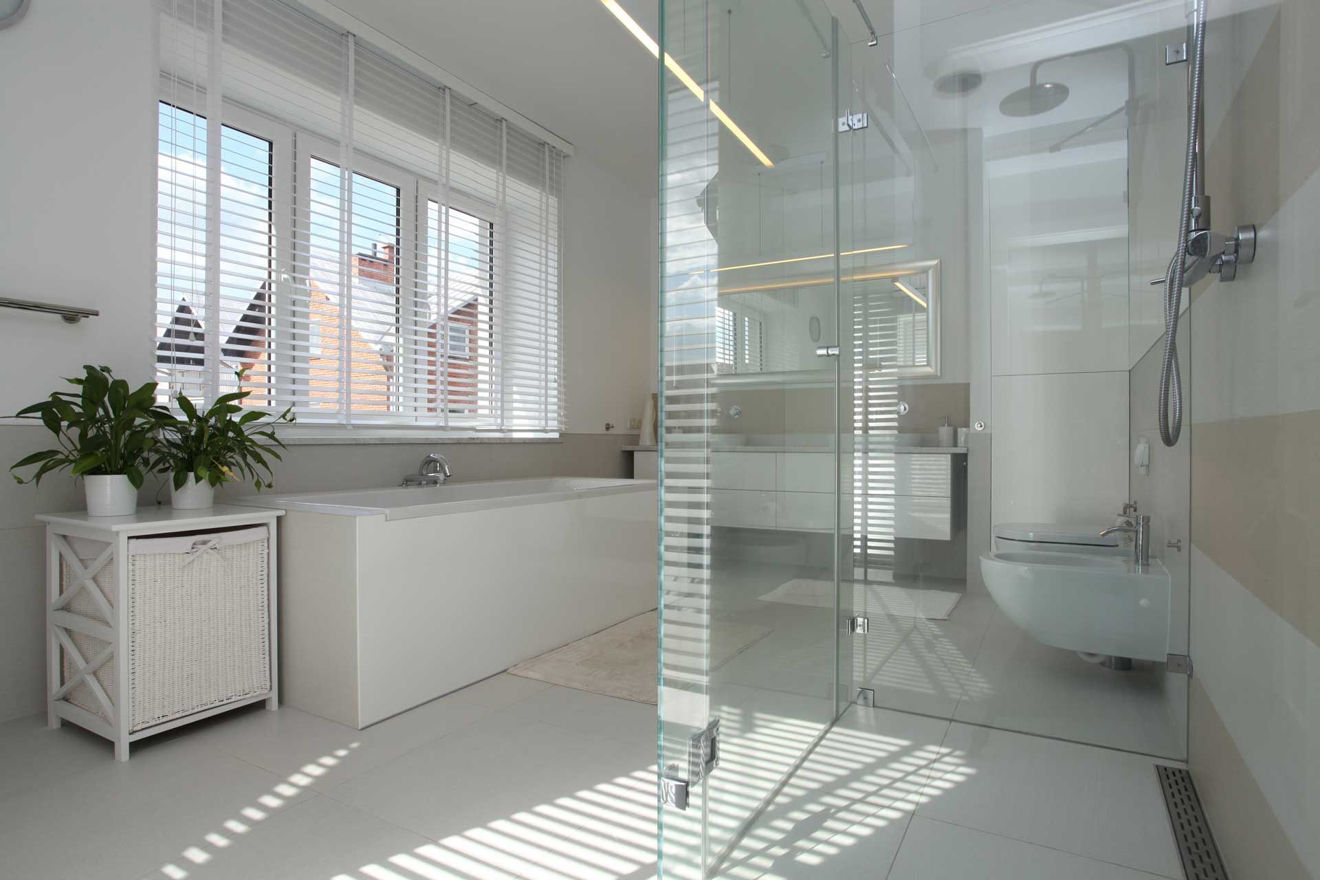Bright, modern bathroom with a glass shower, white tub, and window with blinds.