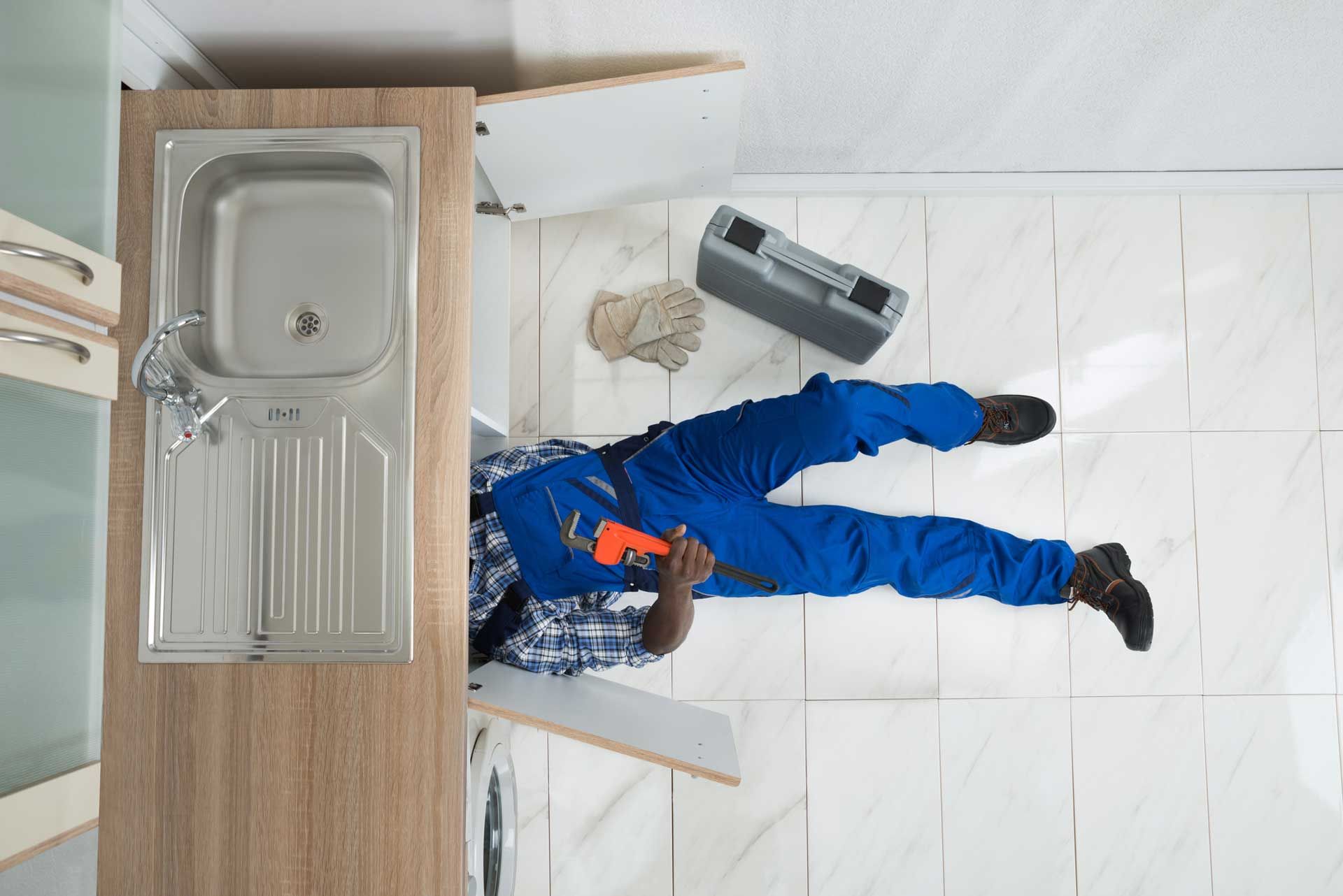 Plumber in blue overalls working under a kitchen sink. The setting is tiled and a radio is present.