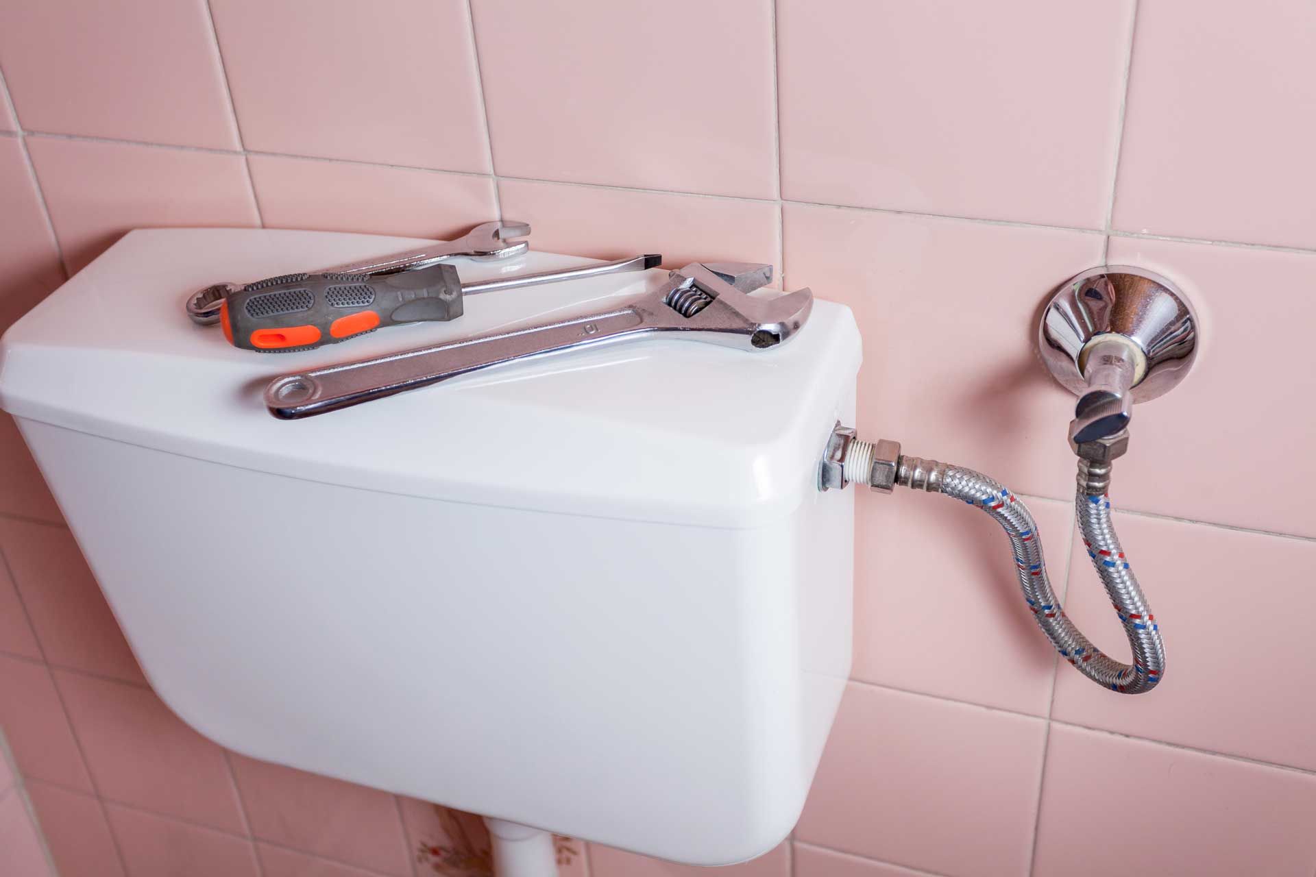 Tools on a toilet tank near a water supply line on a pink tiled wall.