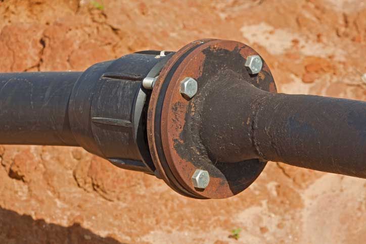 Black pipe sections connected with a rusty flange secured by bolts, set in reddish-brown earth.