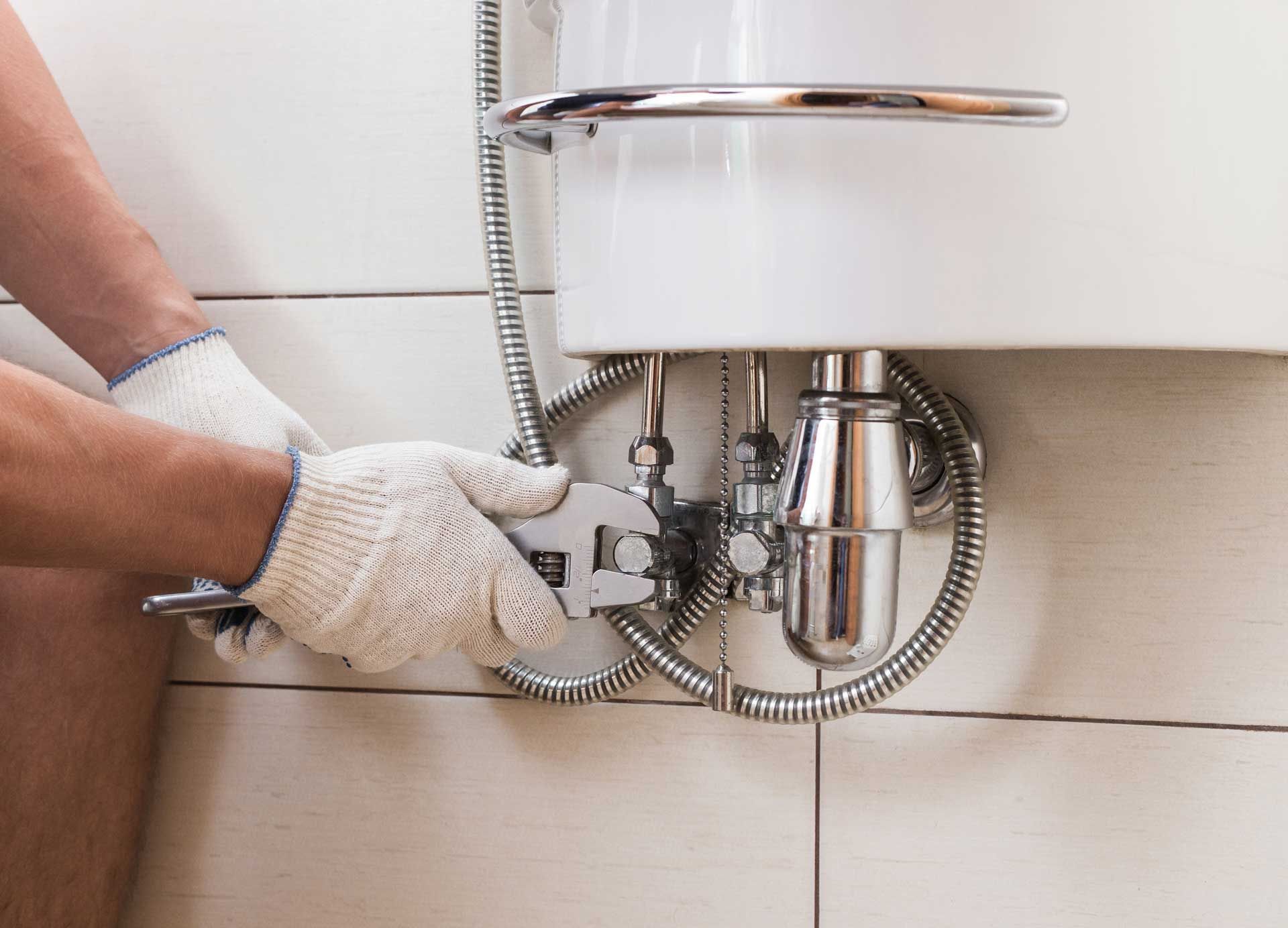 Plumber using a wrench to work on pipes under a white sink in a tiled bathroom.