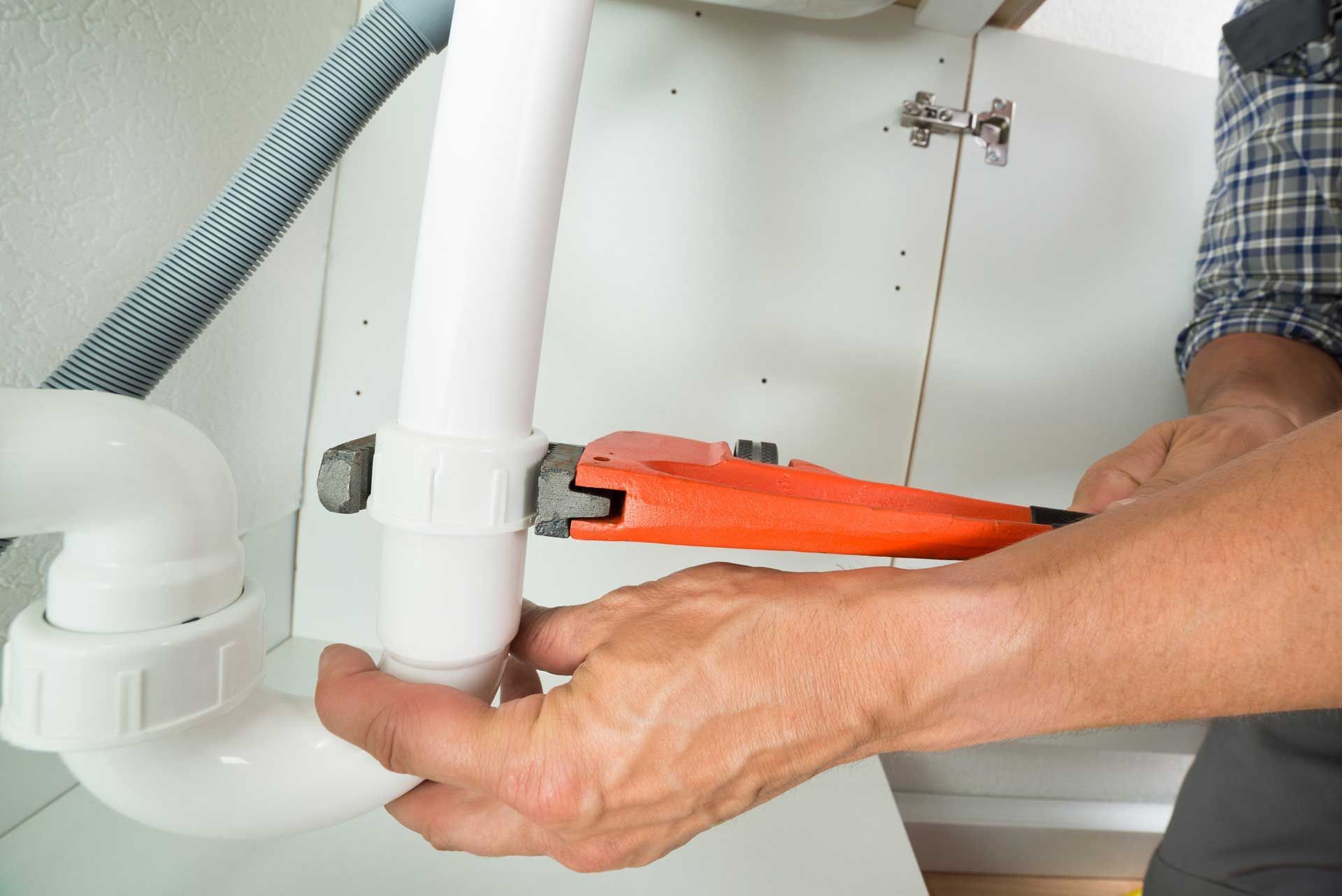 Plumber using a wrench to tighten a white PVC pipe under a sink.