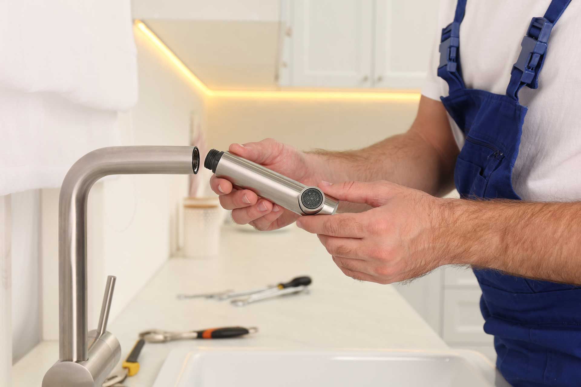Plumber in blue overalls repairing a kitchen faucet, holding a part, with tools on the counter.