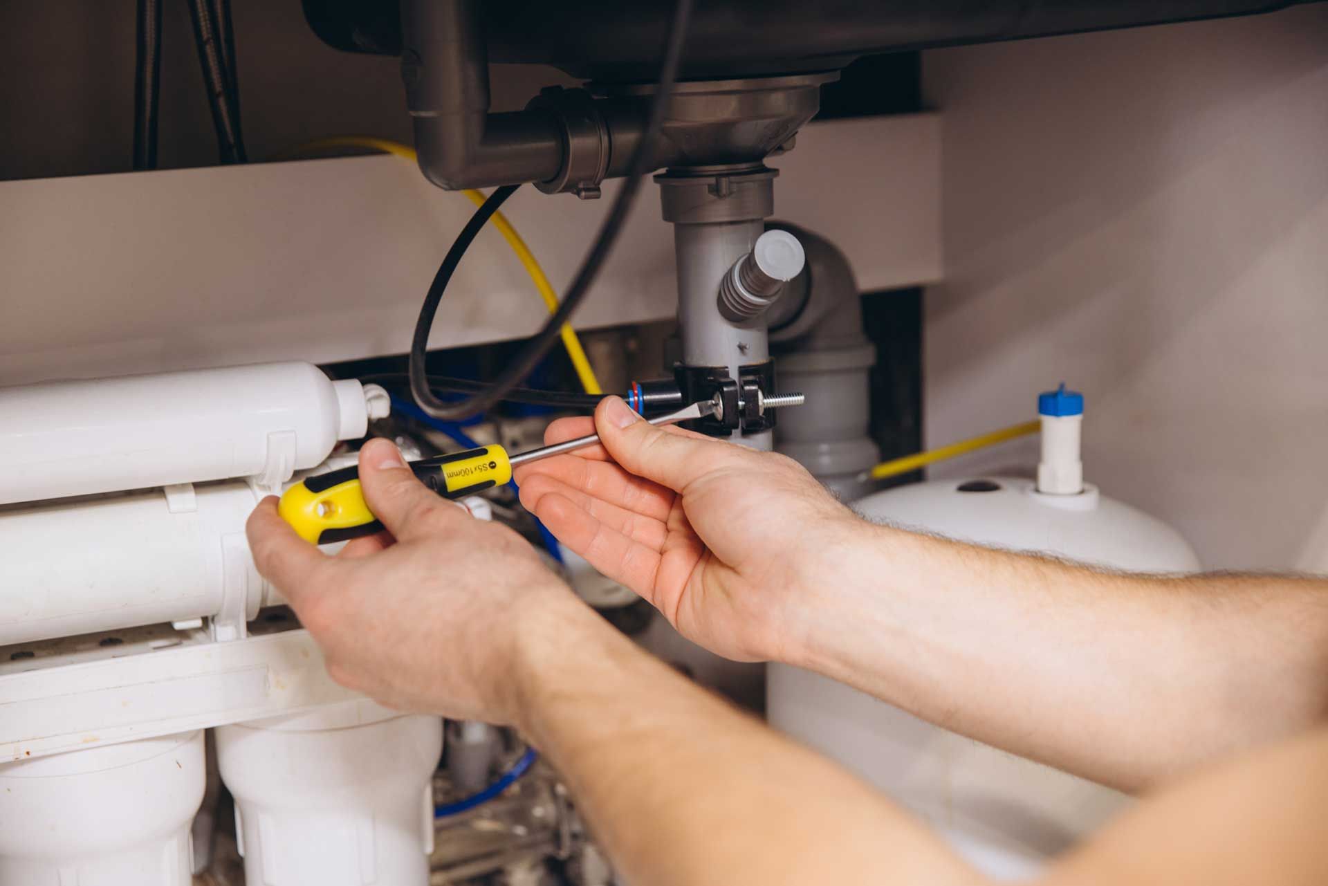 Plumber using a screwdriver to fix pipes under a sink; white plastic and silver metal components.