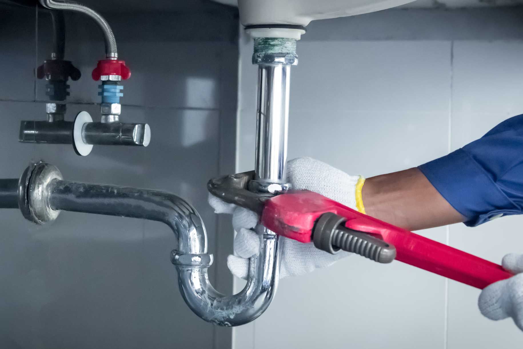 Plumber in blue shirt and white gloves uses a red wrench on a silver drainpipe under a sink.
