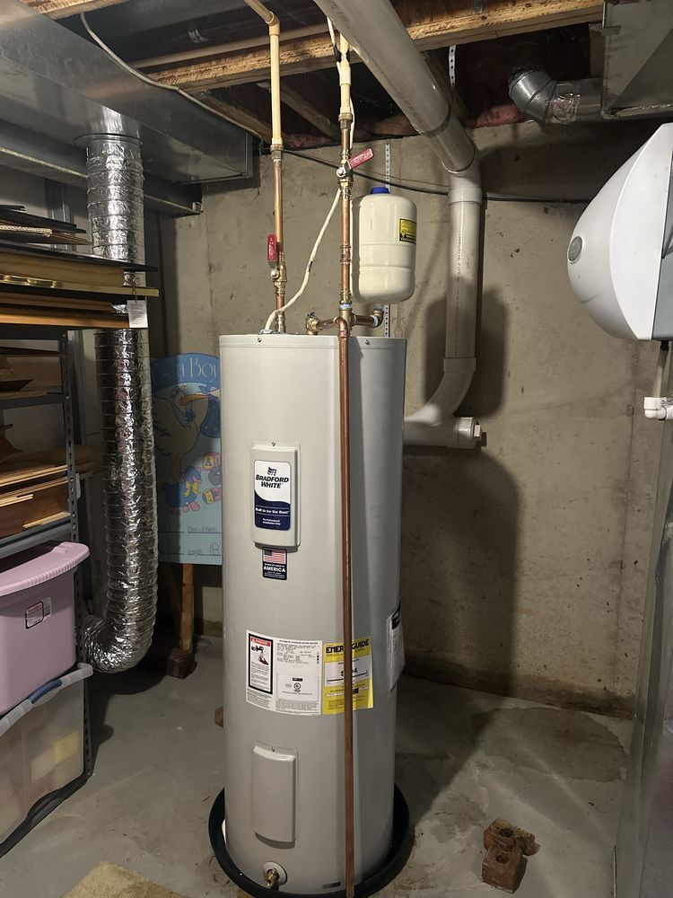 A vertical water heater in a basement. Copper pipes and a small expansion tank are attached to it.