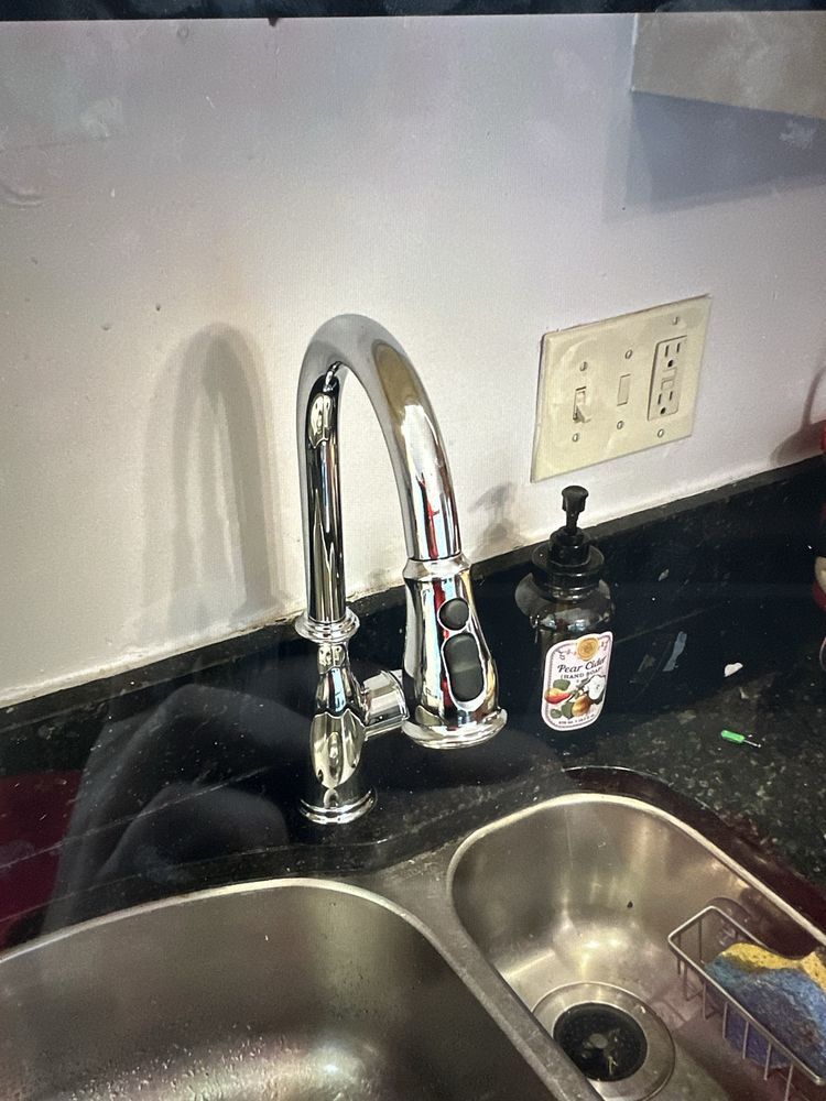 A shiny kitchen faucet over a double sink, with soap dispenser and light switches visible.