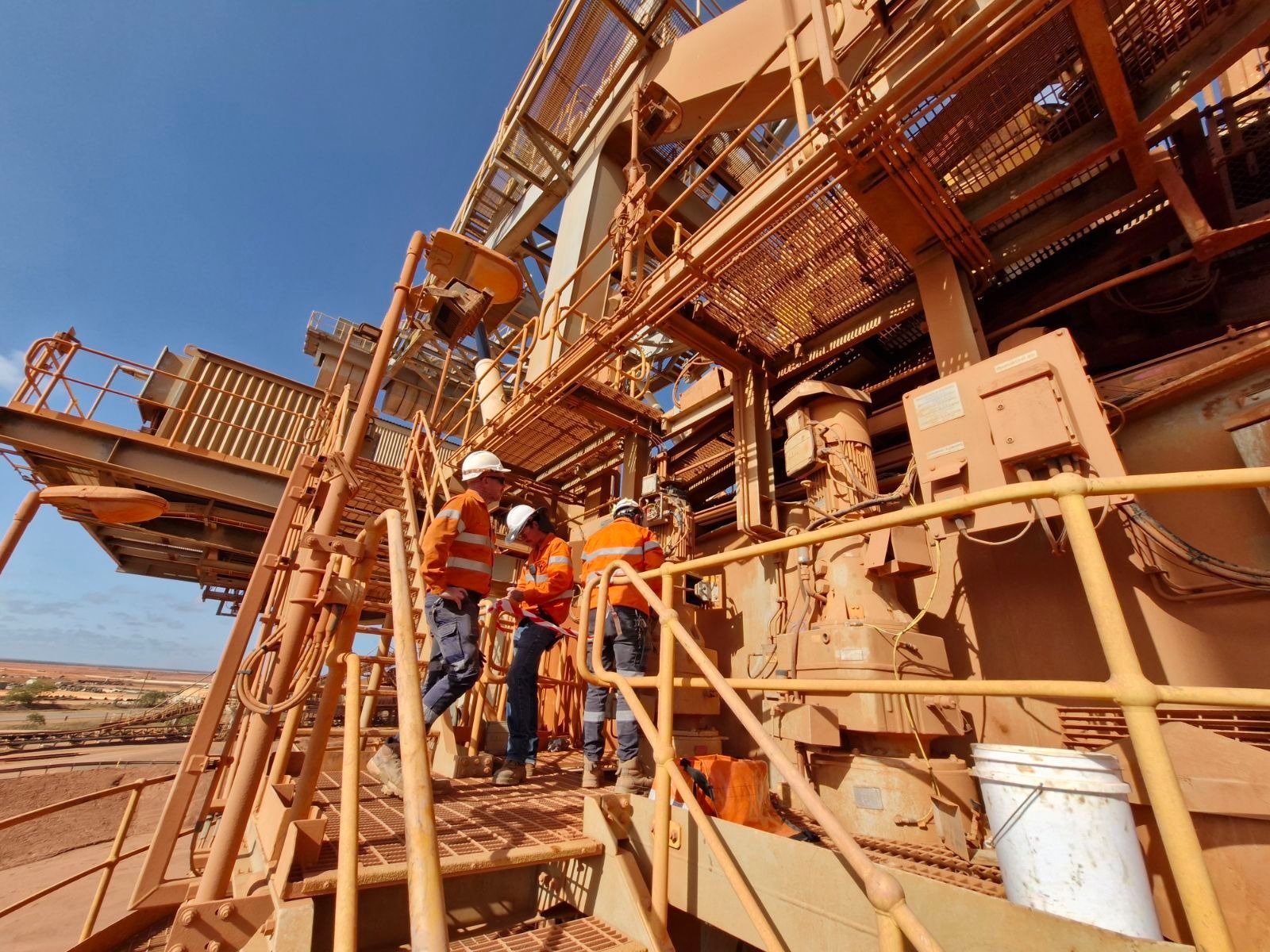 Workers in Orange Vests and Hard Hats Examining Machinery on a Brown Metal Structure Under a Blue Sky — Projected Contracting Electrical Services In Portsmith, QLD