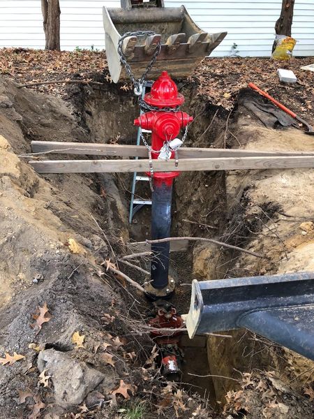 Red fire hydrant in a muddy trench with exposed pipes and wooden planks across it.