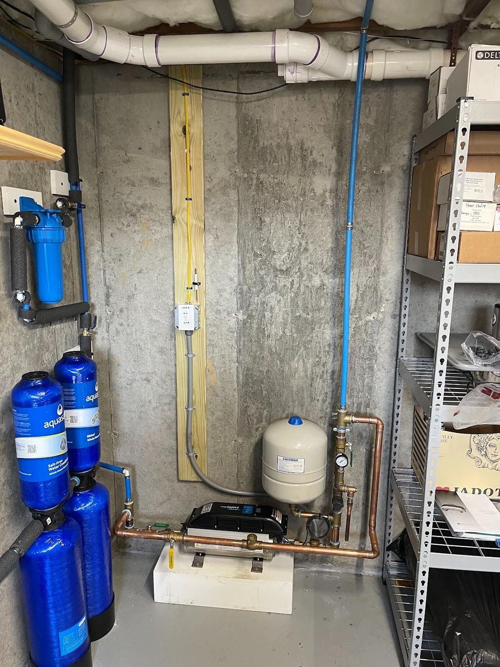 Basement utility area with blue water tanks, pipes, a pump, and shelves against a concrete wall