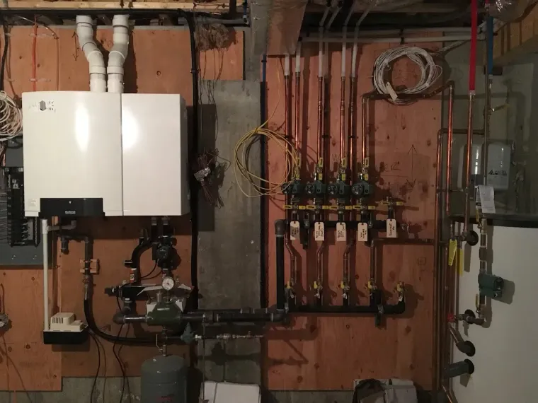 Wall-mounted boiler and plumbing manifold in a utility room with exposed pipes and controls