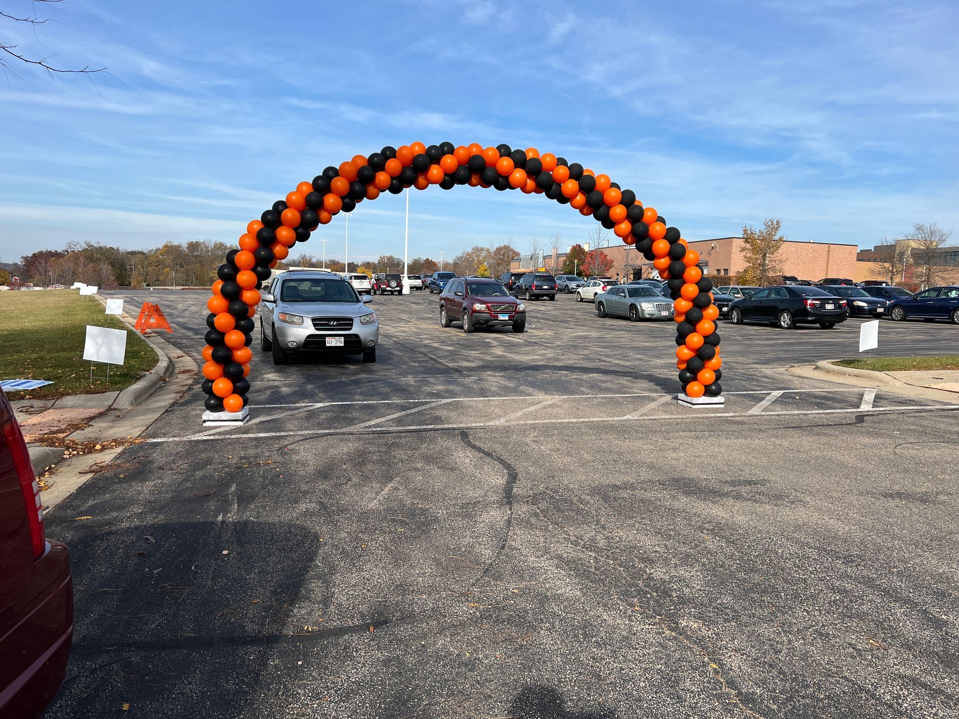 Cars are driving under an orange and black balloon arch in a parking lot.
