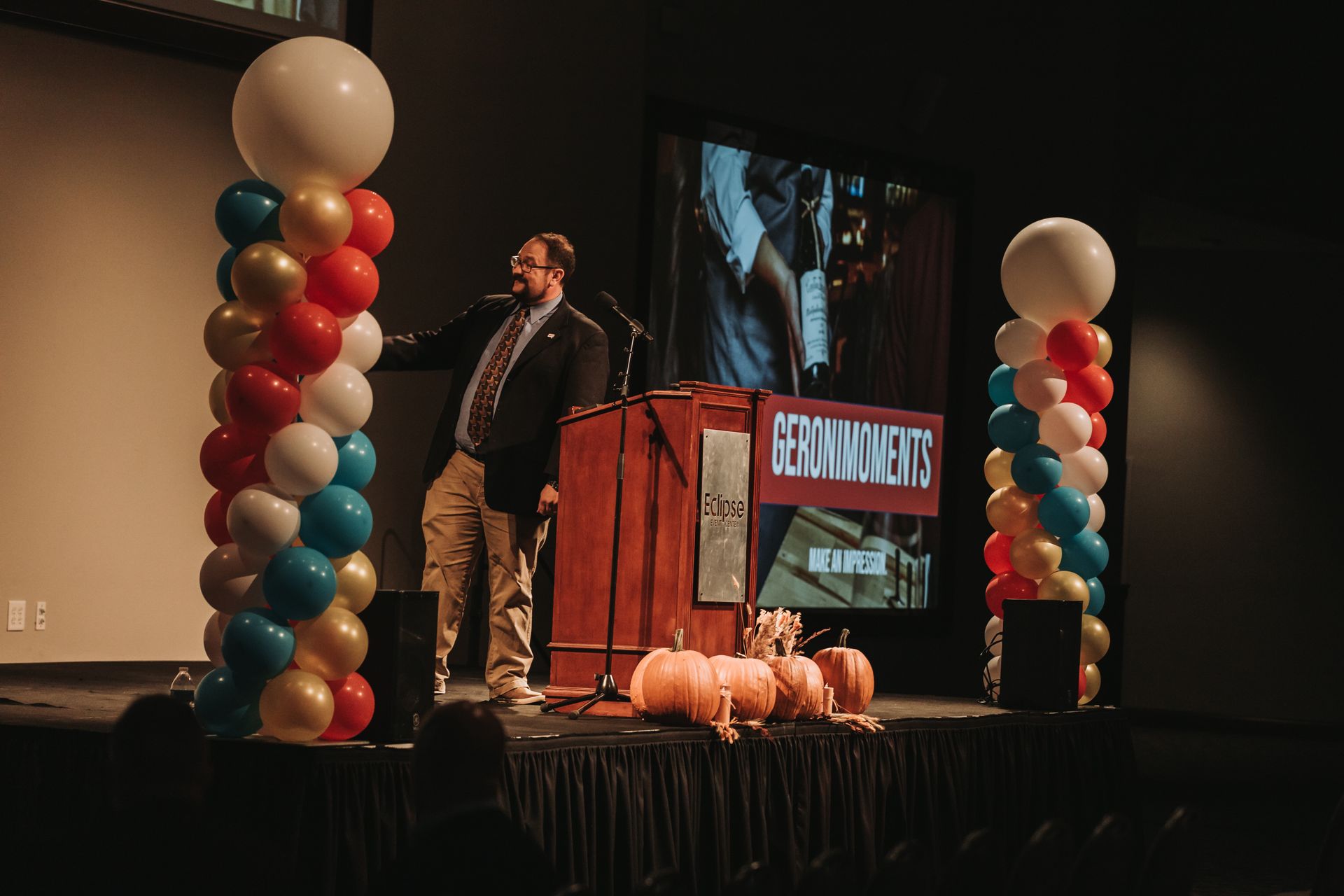 A man is giving a speech on a stage with balloons and pumpkins.