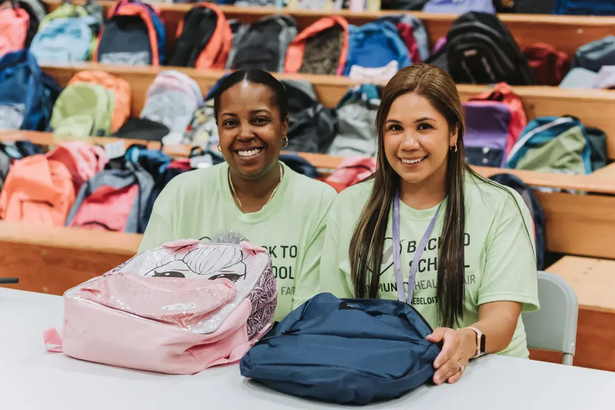 Two women are sitting at a table holding backpacks.
