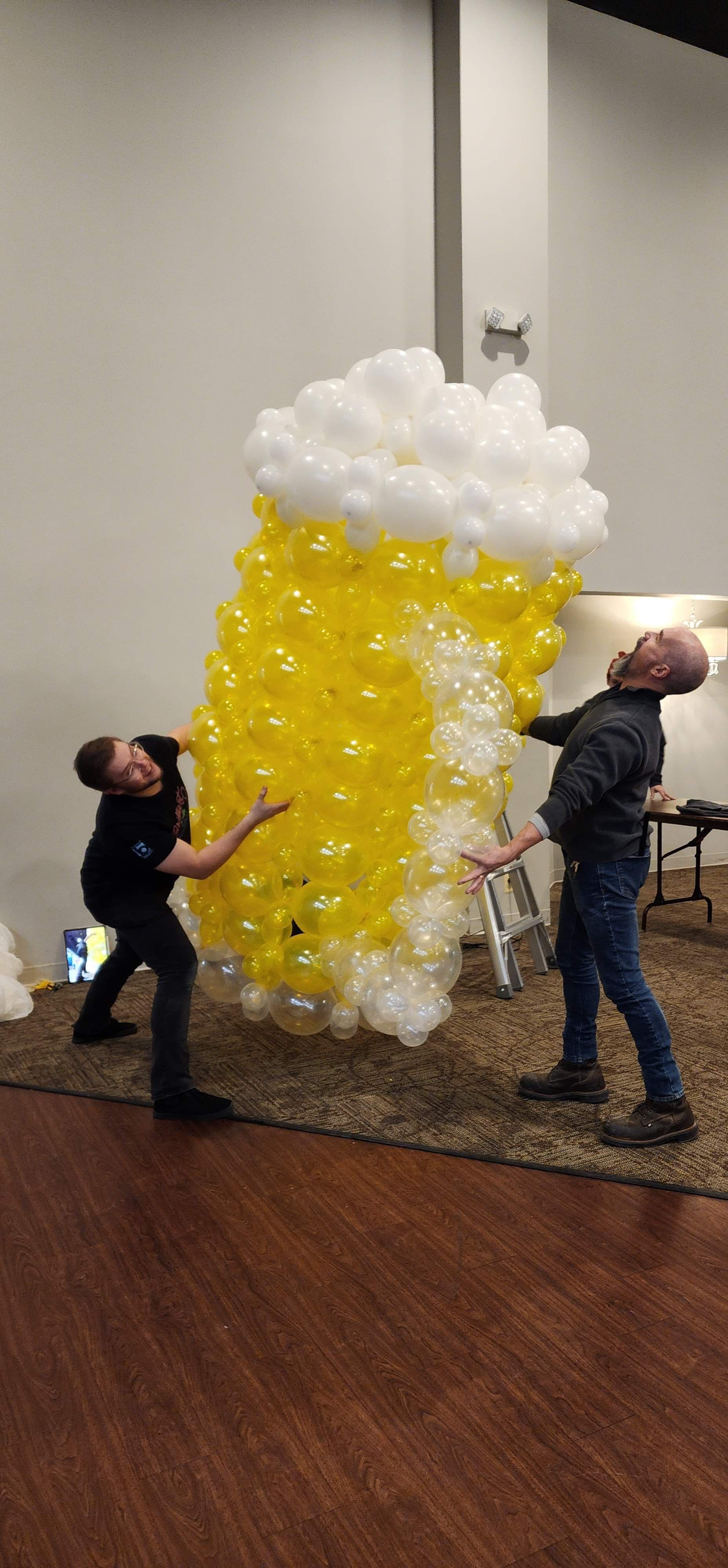 Two men are standing next to a large beer glass made of balloons.
