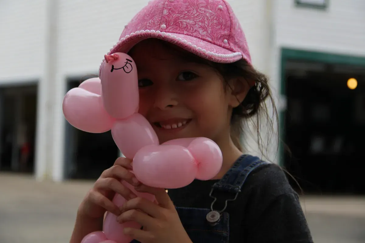 A little girl wearing a pink hat is holding a pink balloon animal.