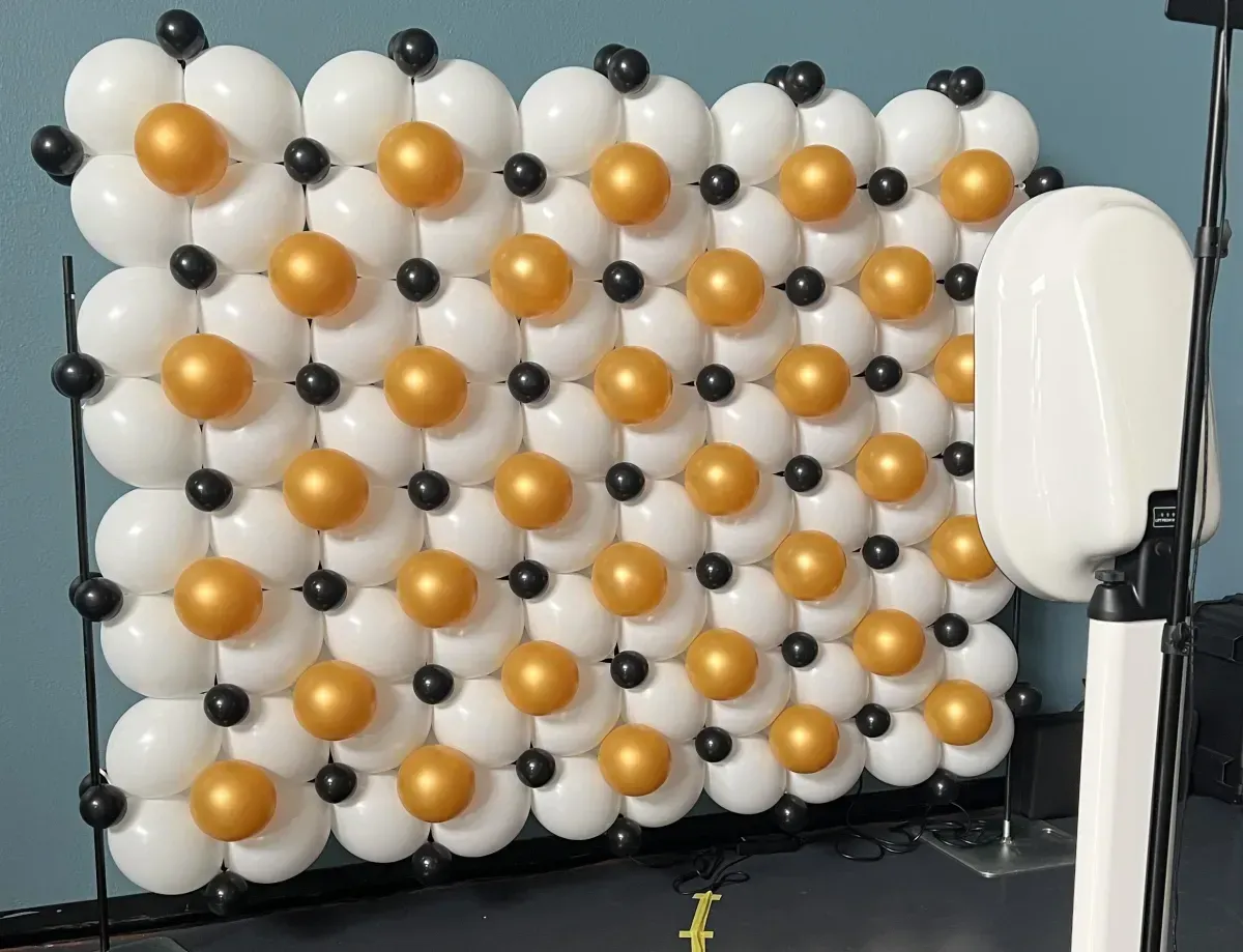 A wall made of white and gold balloons with a photo booth in the background.