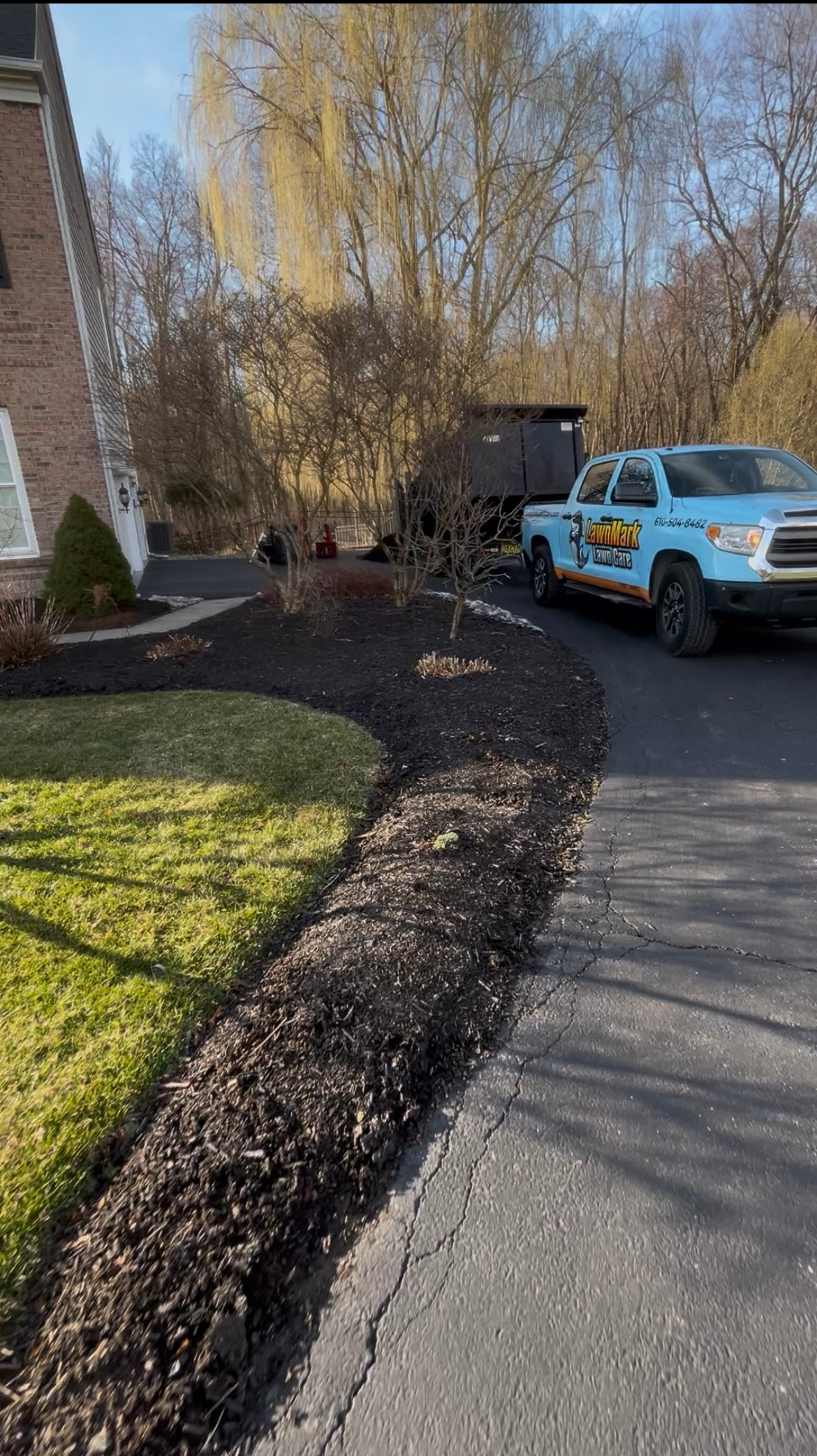 A light blue service truck parked on an asphalt driveway next to a lawn with a fresh layer of dark mulch.