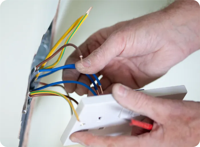 Hands of an electrician wiring a light switch, with visible colored wires and a wall outlet box.