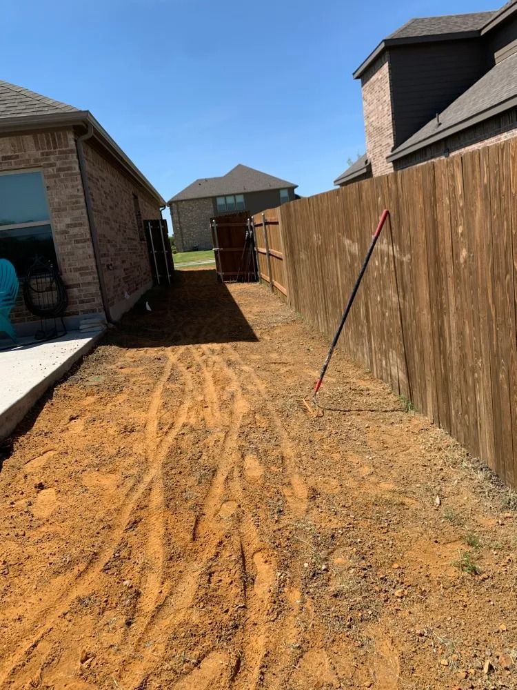 A dirt path between two houses, leading to a gate. Tire tracks mark the ground. A red tool leans against the fence.