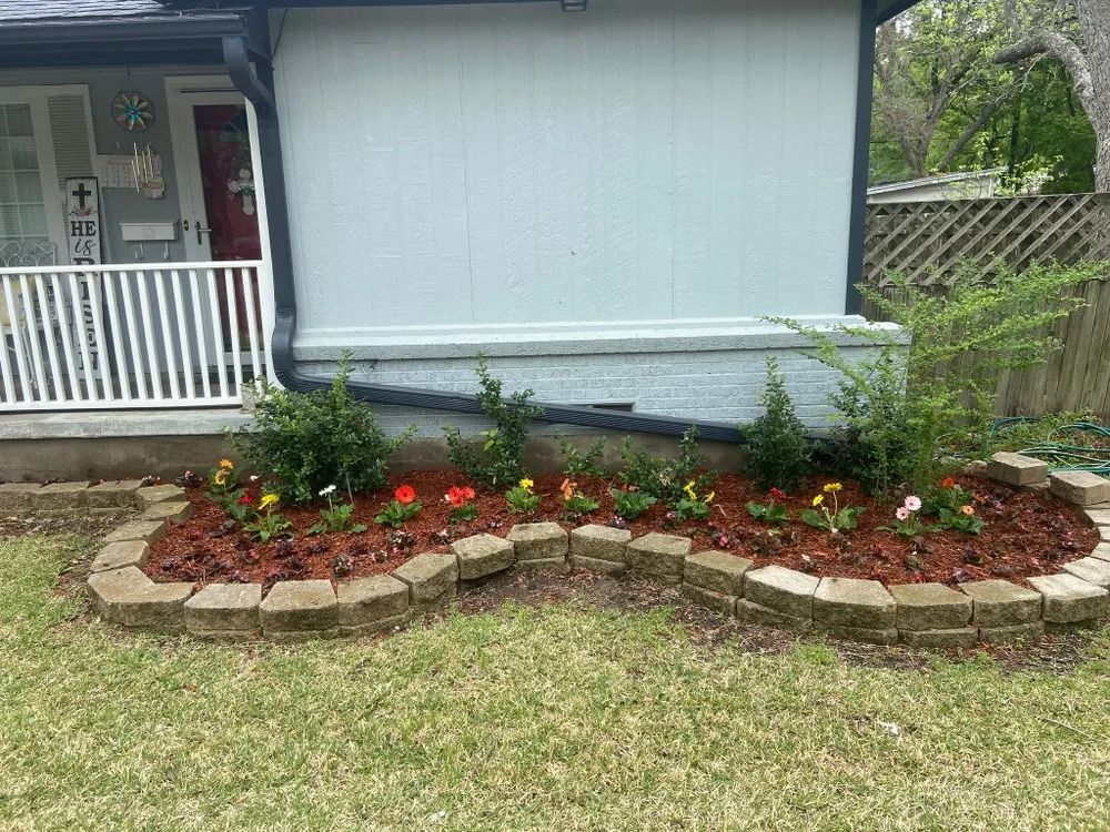 Flower bed in front of a light blue house. Brick edging, mulch, and colorful flowers.
