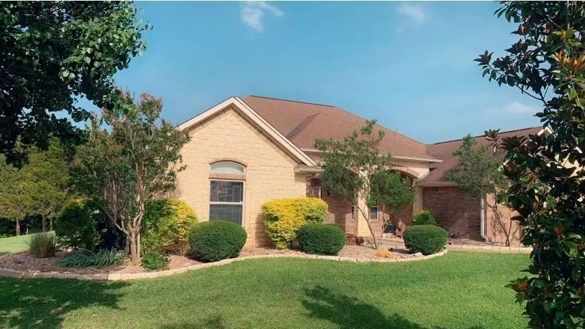 Tan brick house with brown roof, manicured lawn, and landscaping under a blue sky.