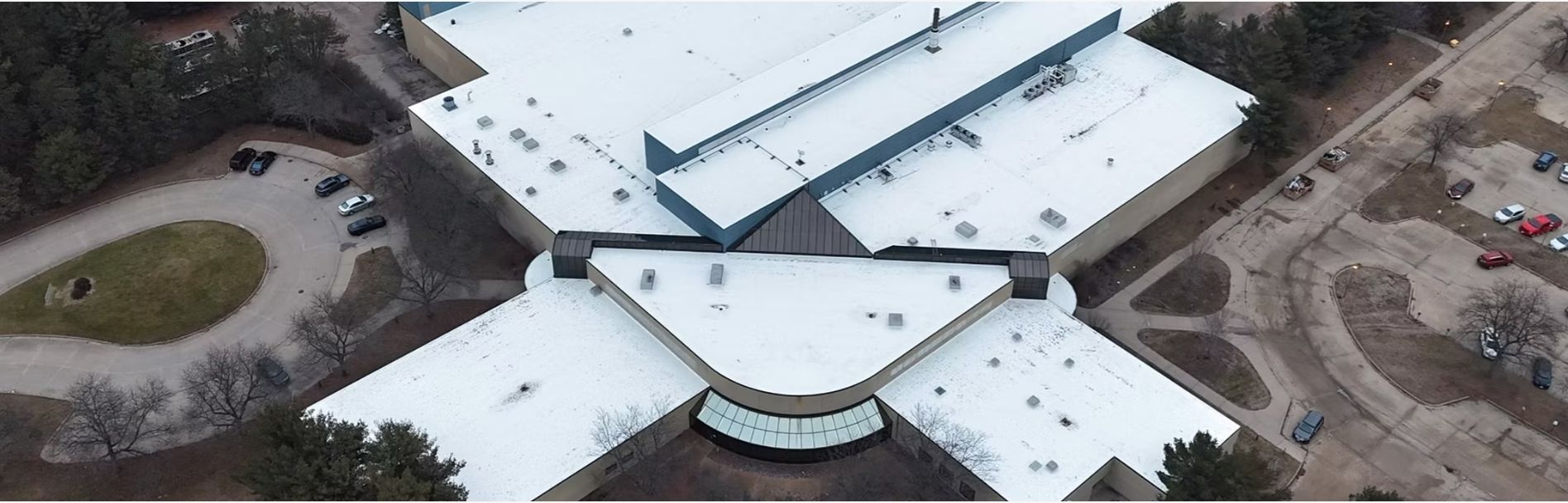 Aerial view of a modern building with a multi-wing design, covered in snow. Parking lots on either side.