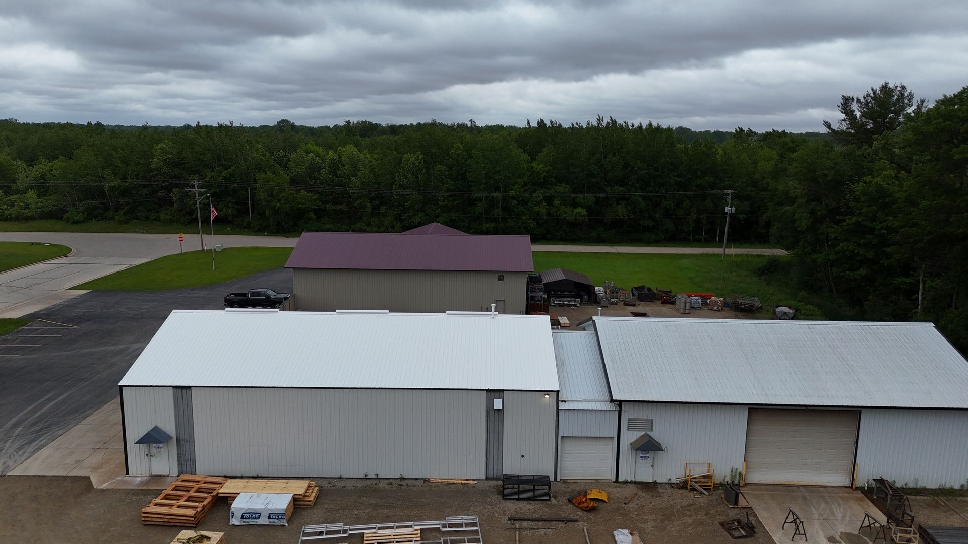 Aerial view of industrial buildings with white and burgundy roofs, trees, and overcast sky.