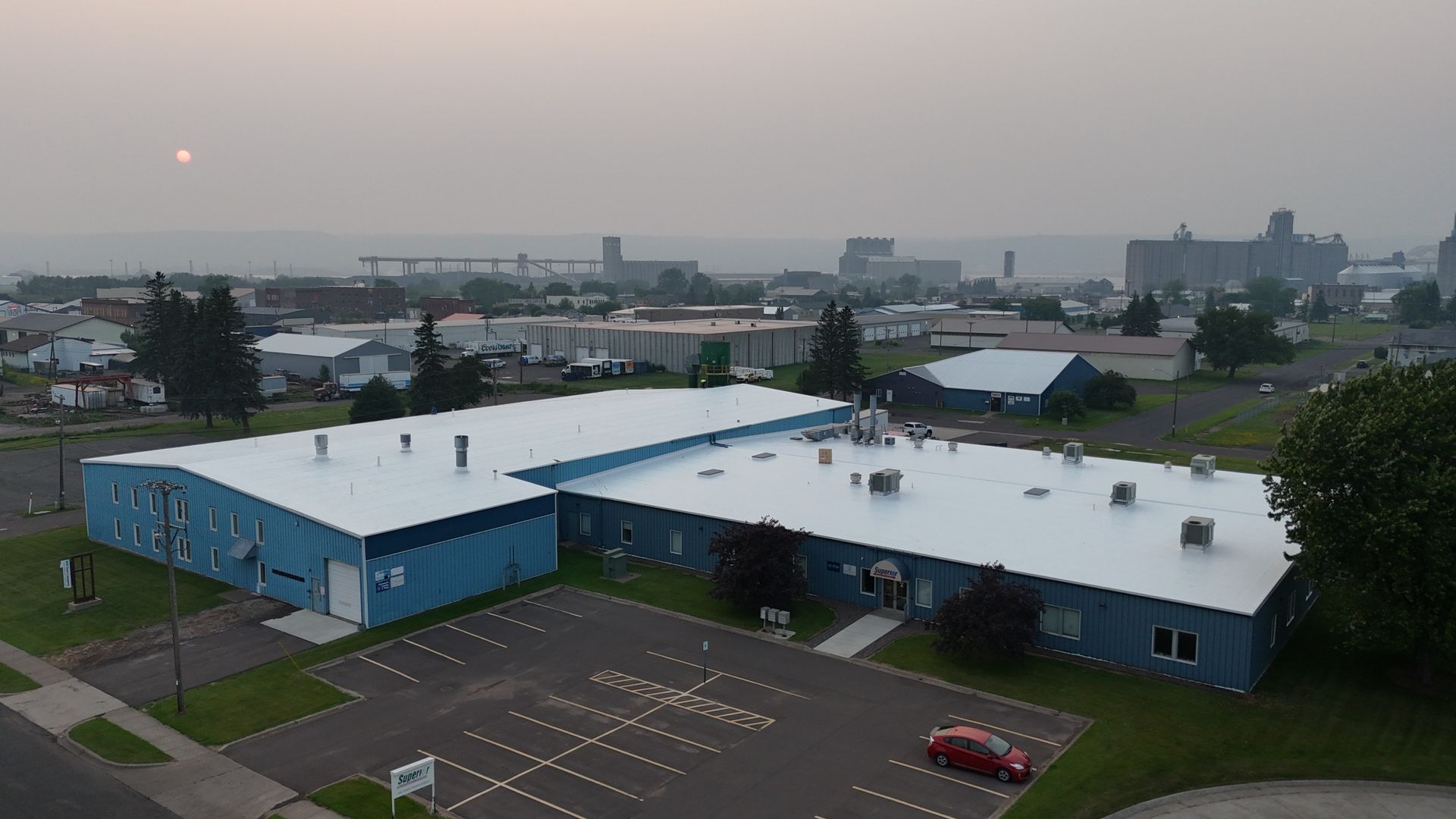 Blue industrial building with white roof, surrounded by a parking lot and other buildings in a hazy, industrial area.