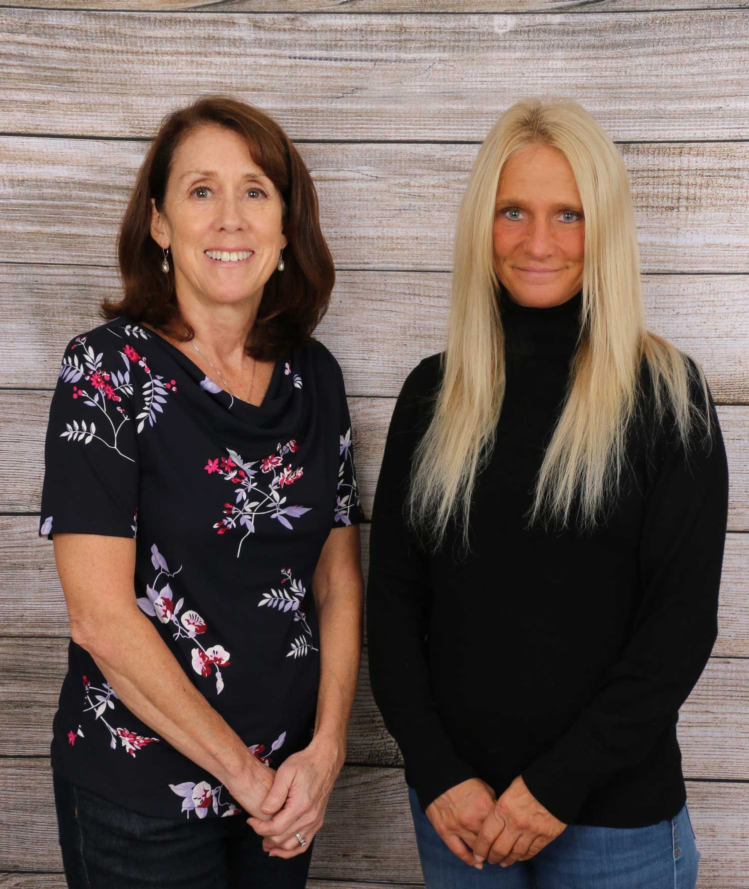 Two women are standing next to each other in front of a wooden wall.